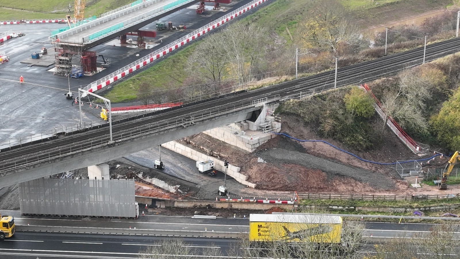 Construction site with bridge, railway tracks, and highway with trucks in daylight.