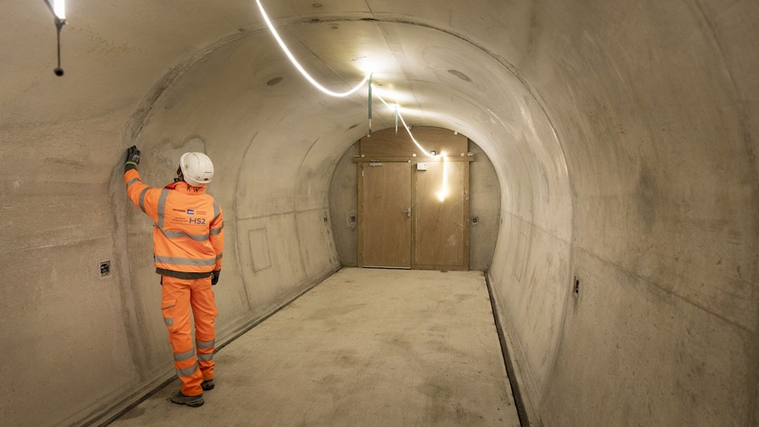 Worker in orange safety gear inspecting tunnel wall under bright overhead lighting.