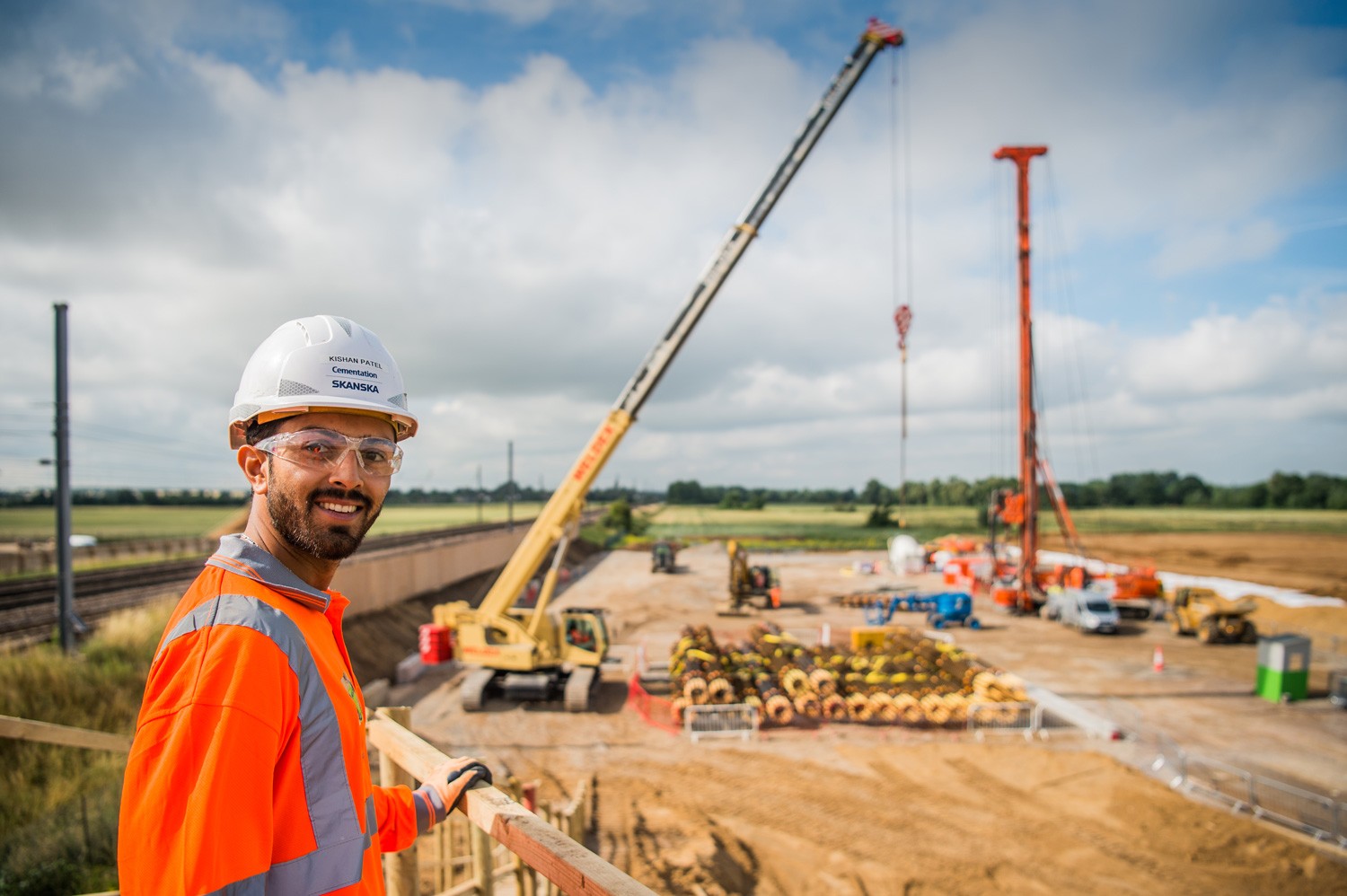 Construction Site with Cranes and Worker in Safety Gear Construction worker oversees site activity, with cranes and materials in a sunny, rural setting.