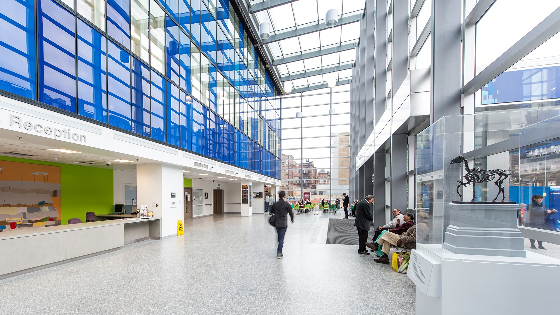 Light and spacious main reception area of the Royal London Hospital Modern hospital lobby with glass ceiling, reception area, and seated visitors waiting.
