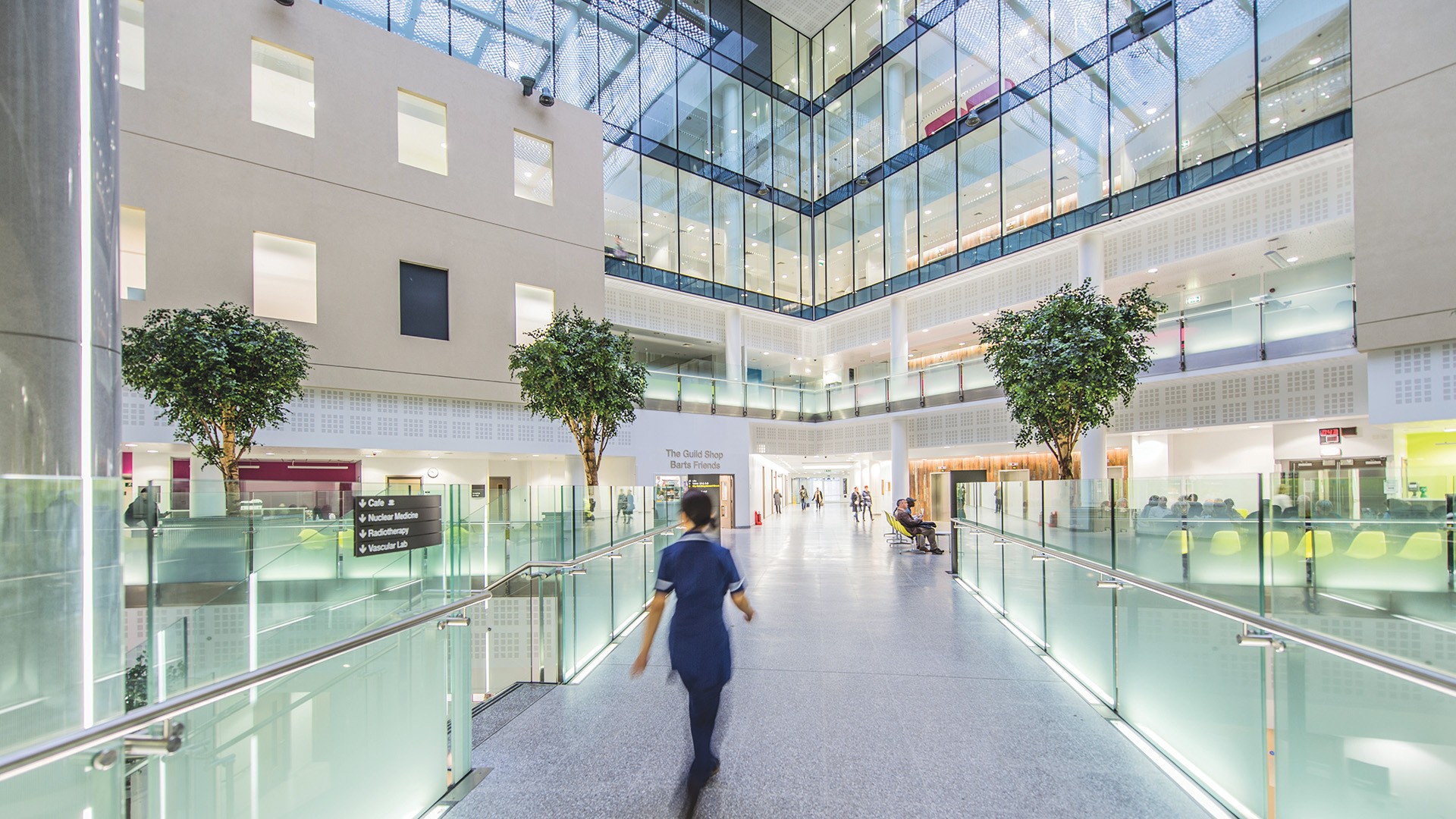 The full height atrium within St Bartholomew’s Hospital Modern hospital atrium with glass walls, large trees, and a nurse walking on a central walkway.