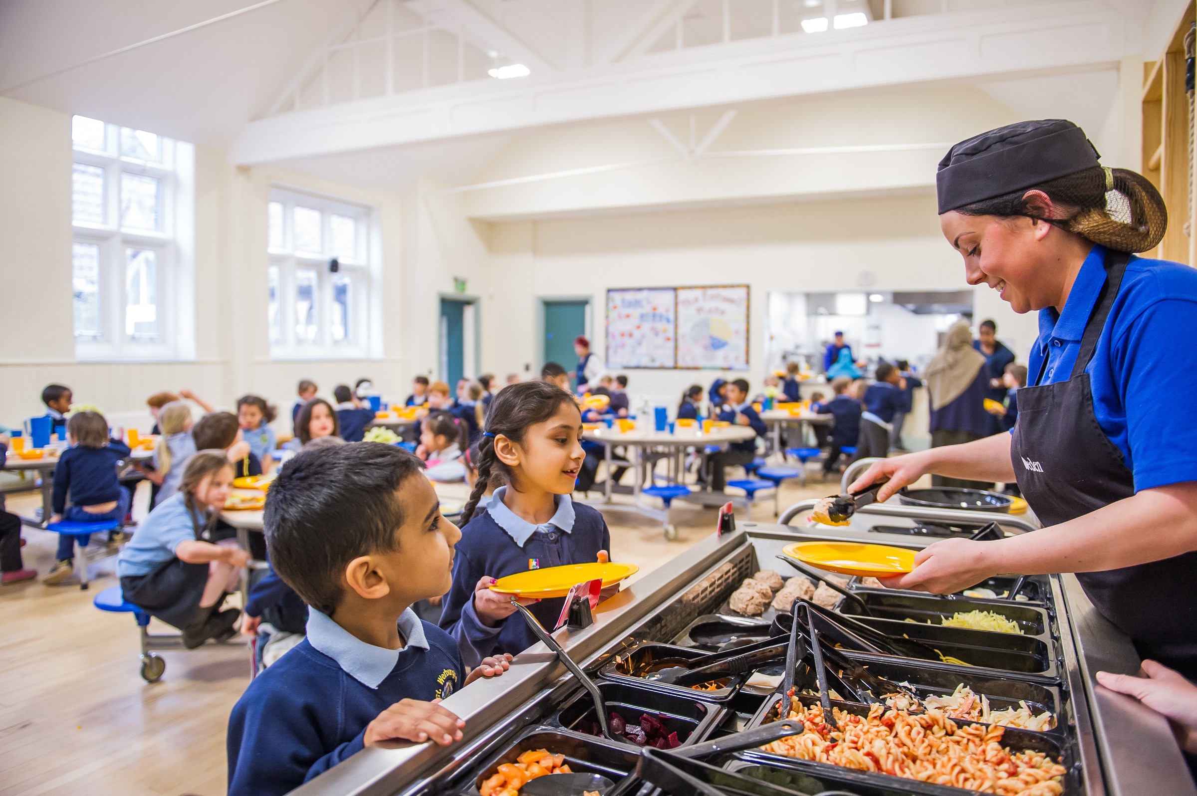 Children in a school cafeteria line up for lunch served from a variety of food trays.