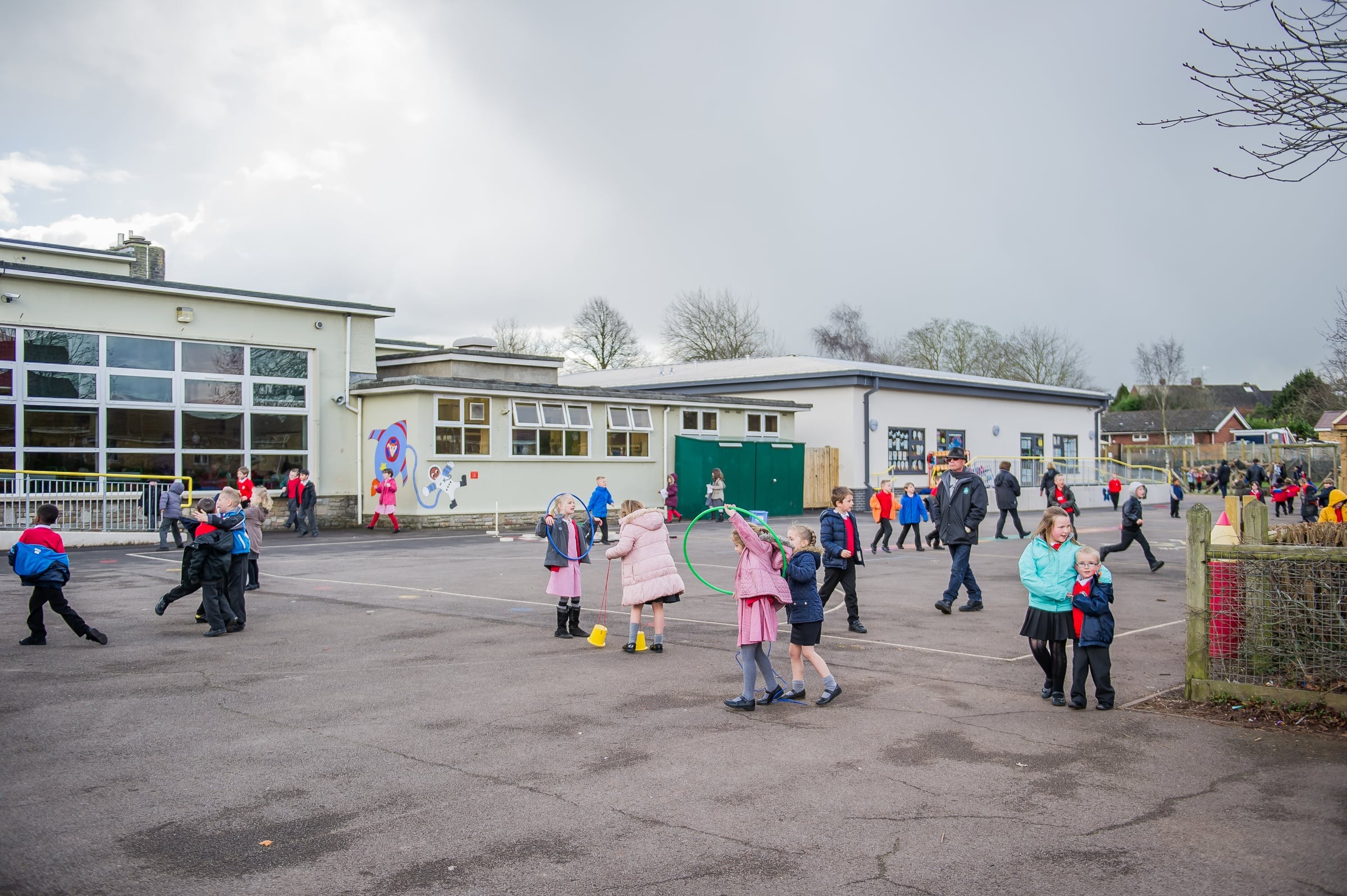Children play in a schoolyard with colorful equipment, surrounded by school buildings and trees.