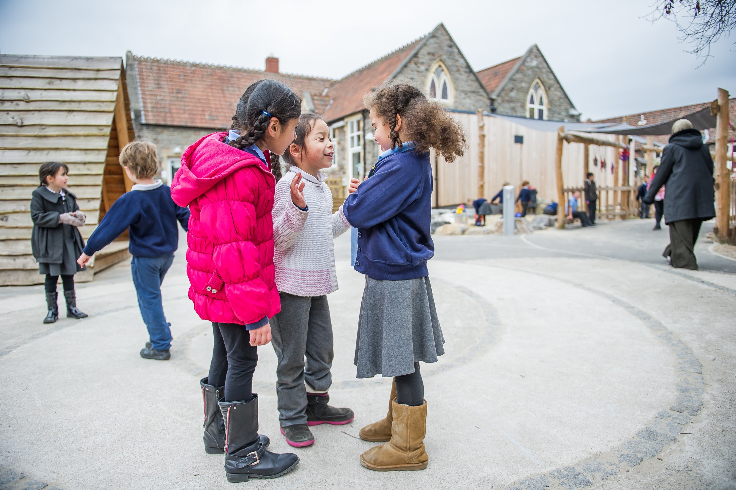 Playtime fun in St Werburghs Primary’s landscaped exterior space Children interacting in a school playground with a stone building in the background.