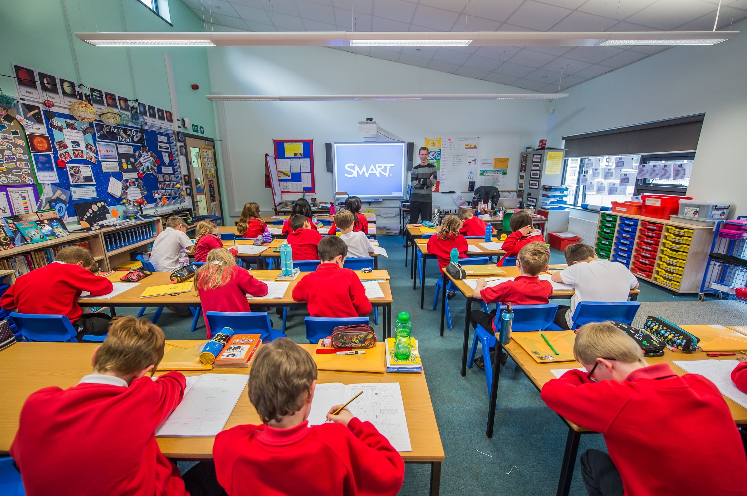 Students in red uniforms focus on a lesson in a classroom with a SMART board presentation.