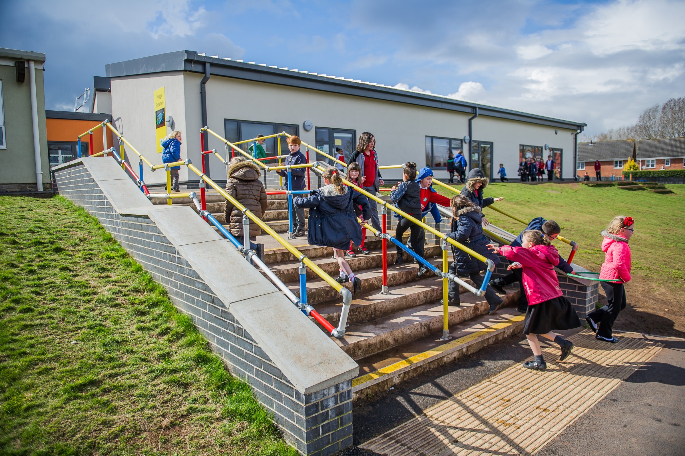 Children playing on colorful stairs outside a modern school building under a partly cloudy sky.