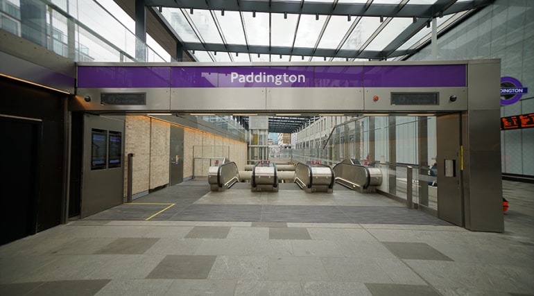 Entrance to Paddington Station with escalators and a purple overhead sign under a glass roof.