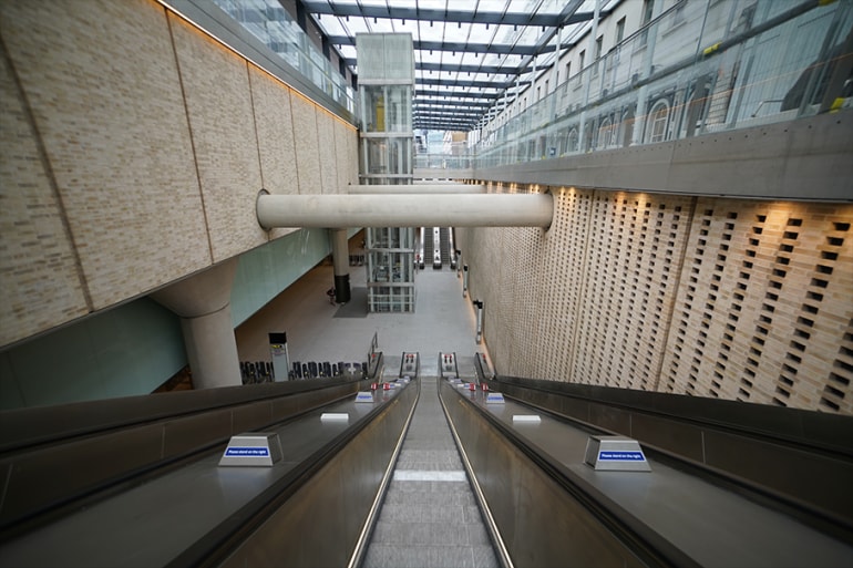 Modern escalators descend into a spacious, glass-roofed atrium with brick walls and an elevator.