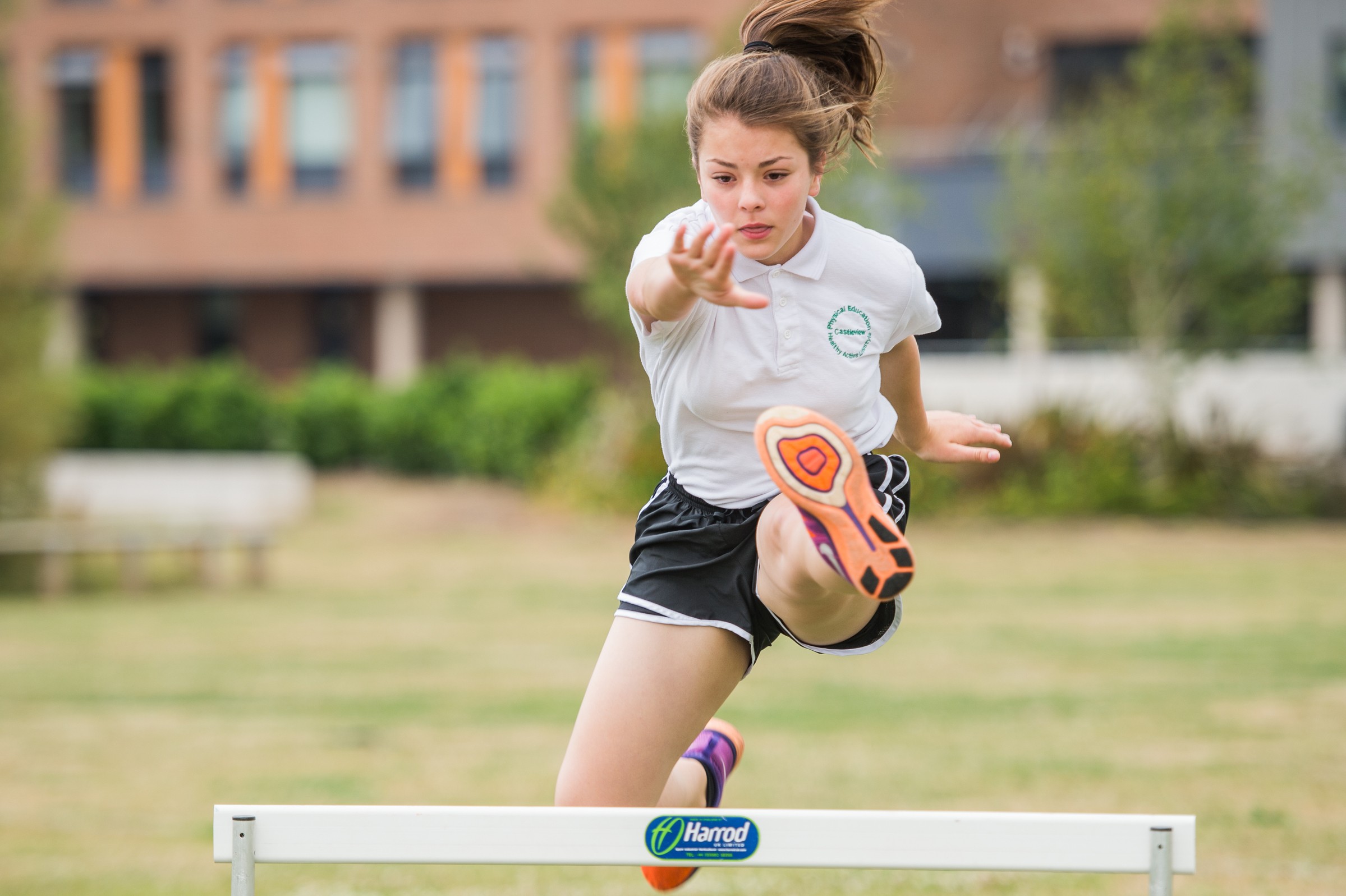 A young athlete in a white shirt and black shorts hurdles over a Harrod-branded barrier outdoors.