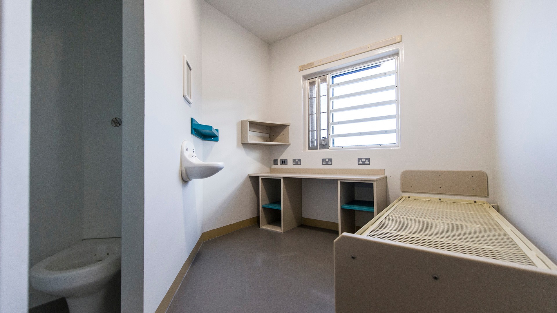 Minimalist prison cell with barred window, single bed, toilet, sink, and built-in desk unit.
