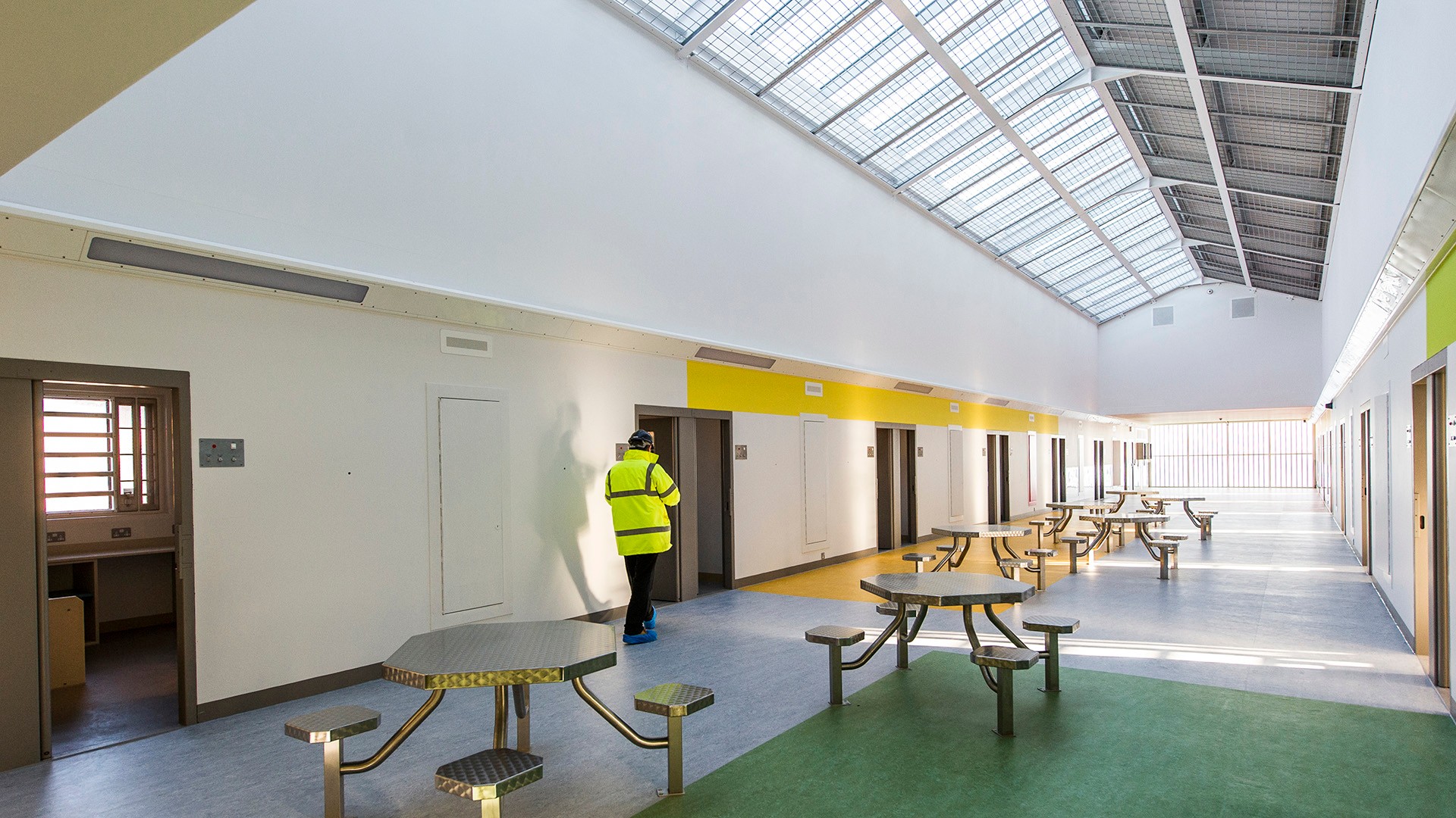 Modern prison interior with skylights, metal tables, and a person in high-visibility clothing.