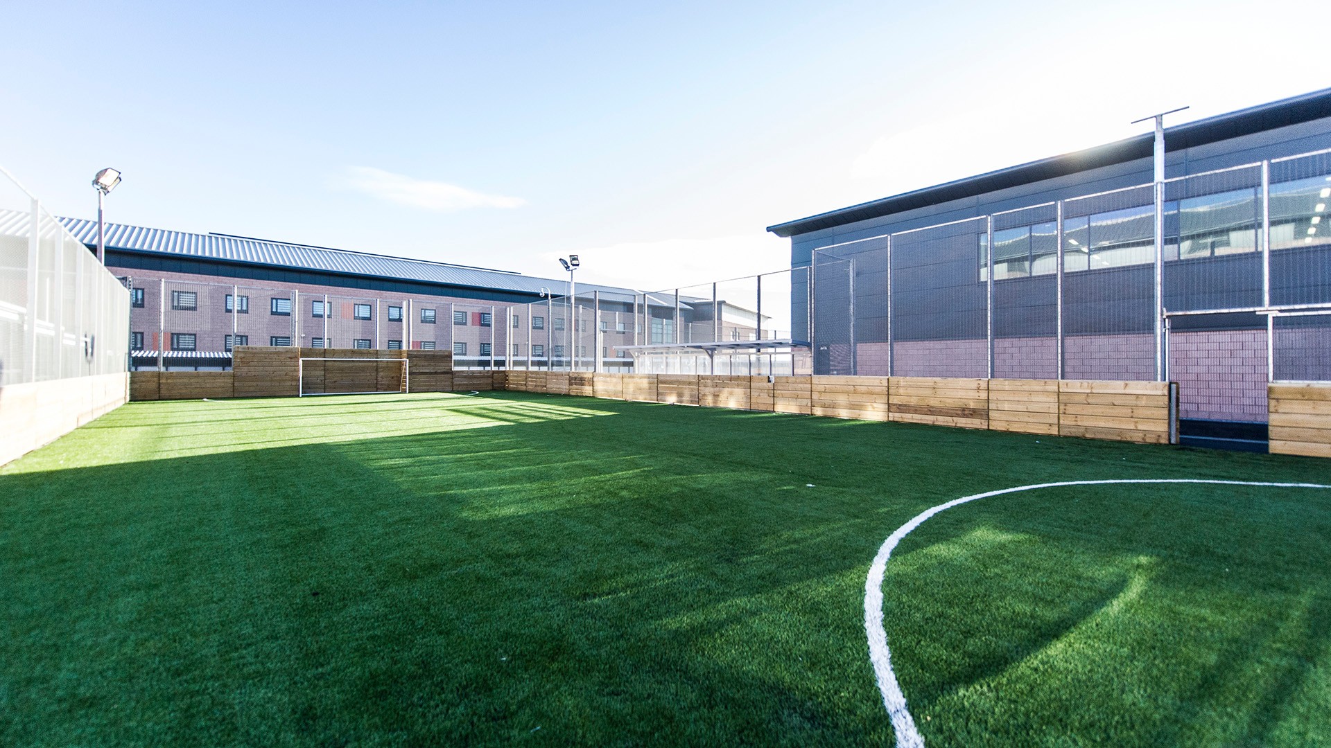 Outdoor artificial turf soccer field surrounded by buildings and protective fencing.