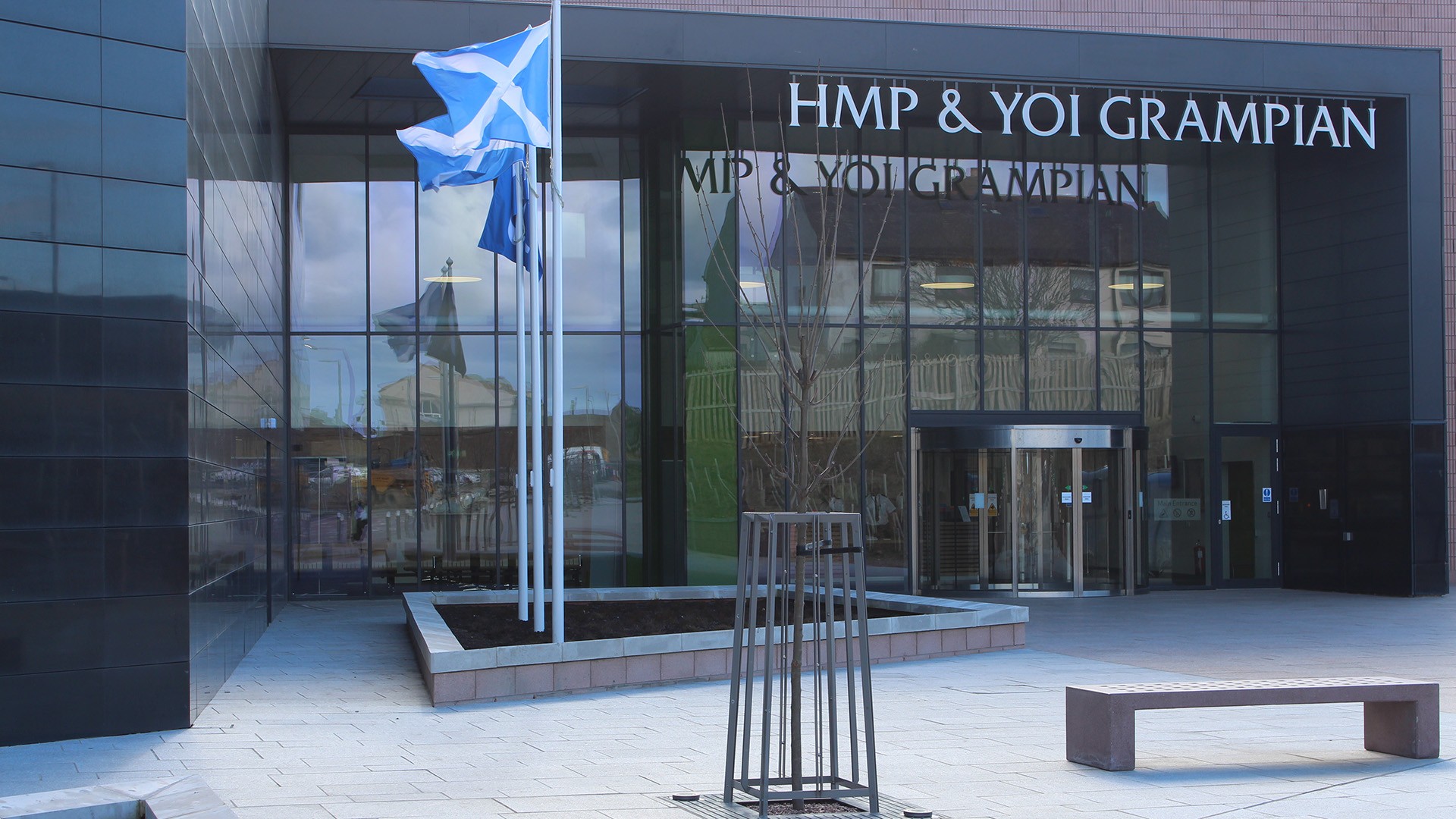Entrance of HMP & YOI Grampian with Scottish flags, featuring modern architecture and large glass doors.