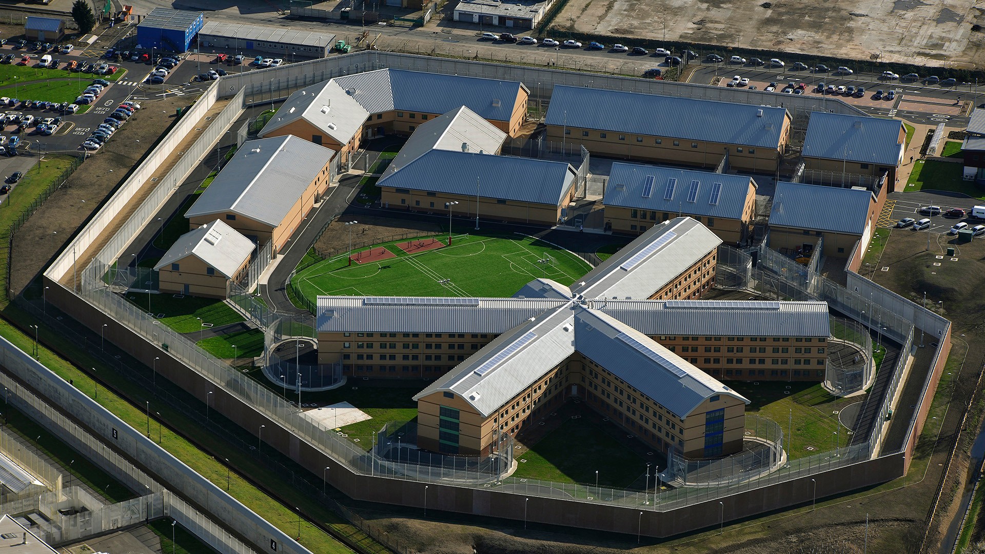 Aerial view of a modern prison complex with X-shaped buildings, surrounded by high security fences.
