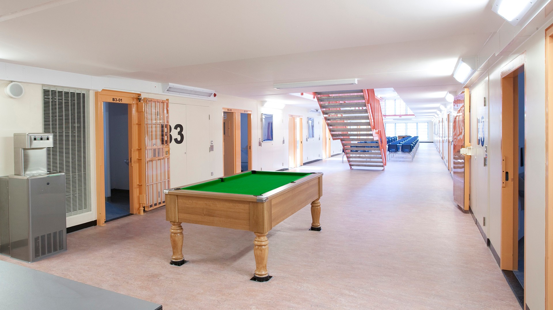Modern prison interior with a pool table, metal staircase, and numbered cell doors in a hallway.