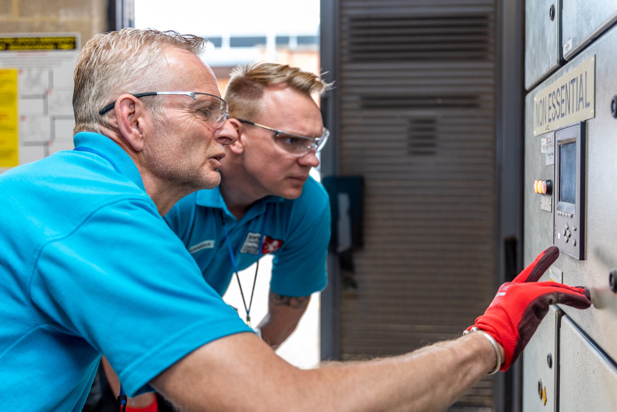 Two technicians in blue uniforms operate a control panel labeled "NON ESSENTIAL" with focus.