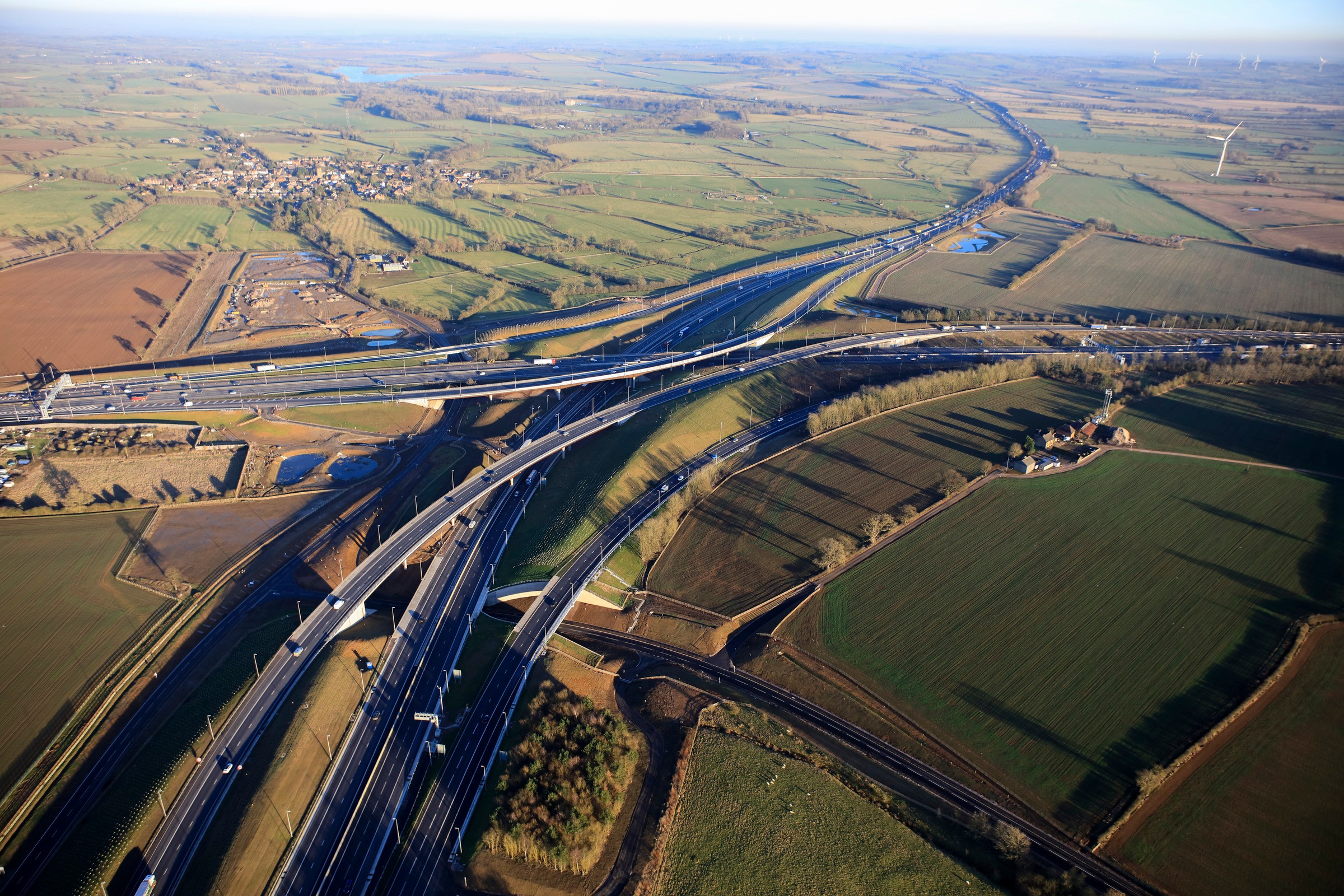 M1 Junction 19 looking east Aerial view of a complex highway interchange surrounded by lush green fields and rural landscape.