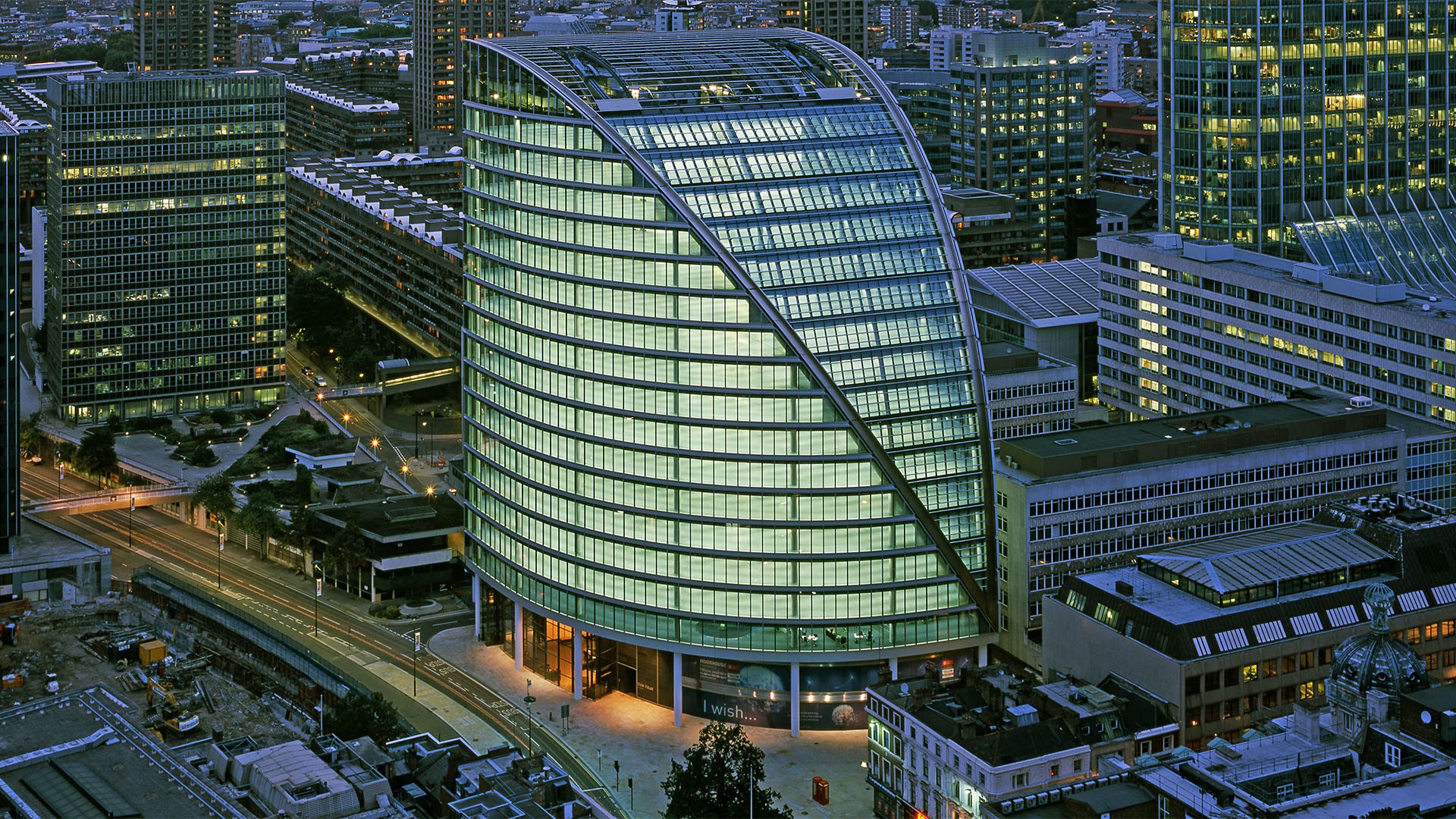 Modernes Bürogebäude mit gebogenem Glas in städtischer Umgebung, umgeben von Stadtlandschaft in der Dämmerung.