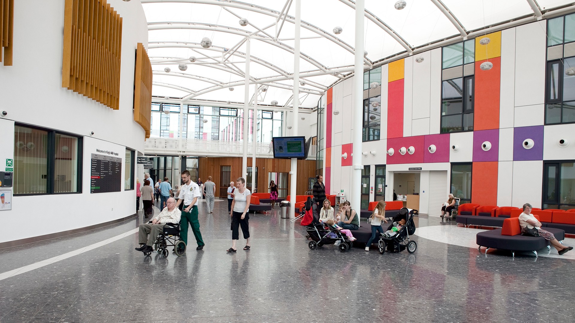 Modern hospital lobby with colorful walls, people in wheelchairs, and visitors sitting or walking.