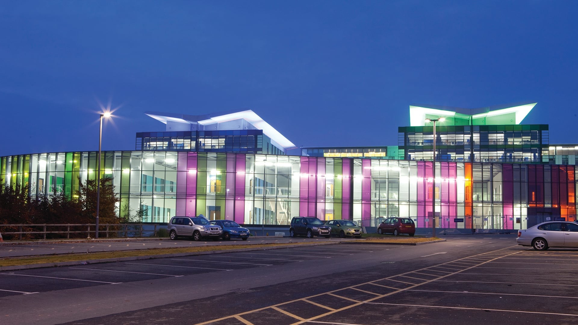 Colorful, illuminated modern building at night with cars parked in the foreground.