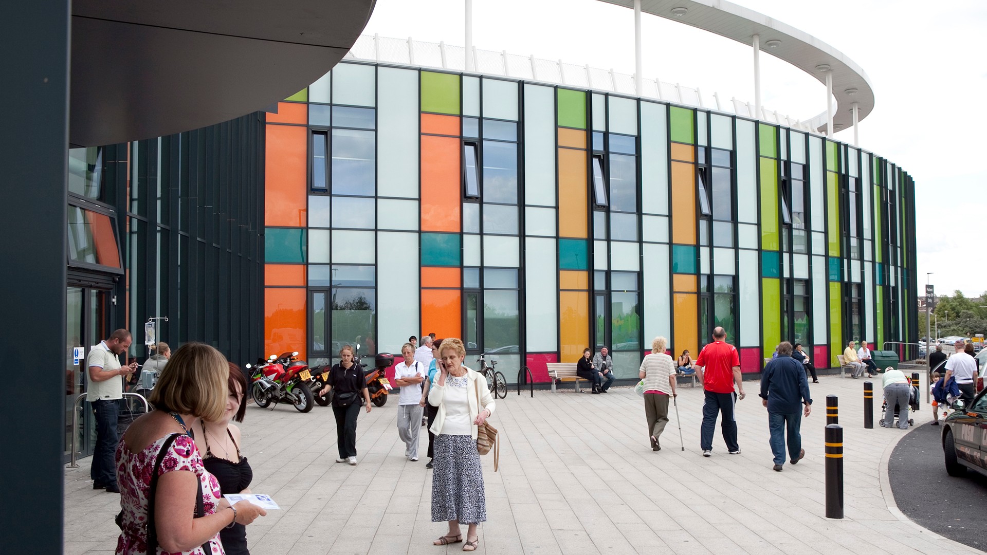 Modern building with colorful glass facade and people walking outside on paved area.