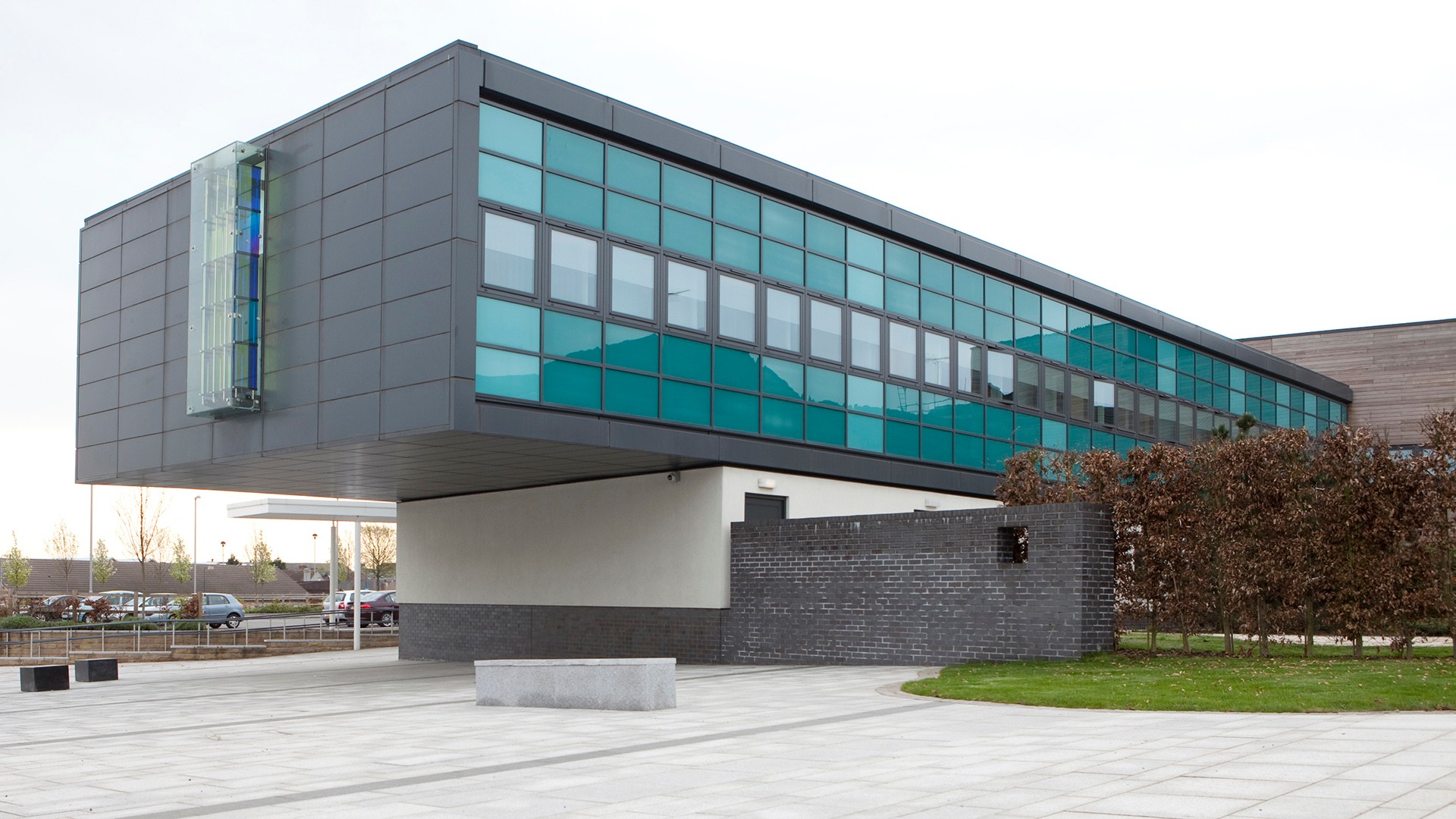 Modern building with teal glass windows, overhanging design, and landscaped surroundings.