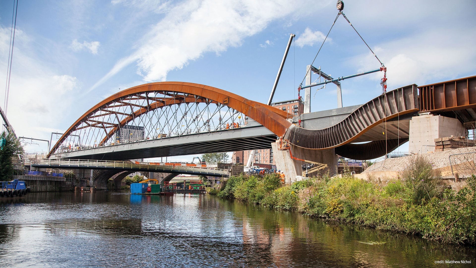 Construction of a modern arched bridge over a river with cranes and workers in an urban setting.