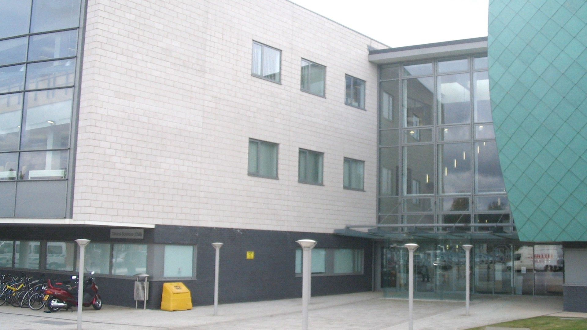 Modern building facade with glass entrance, yellow bin, and bicycles; overcast sky backdrop.