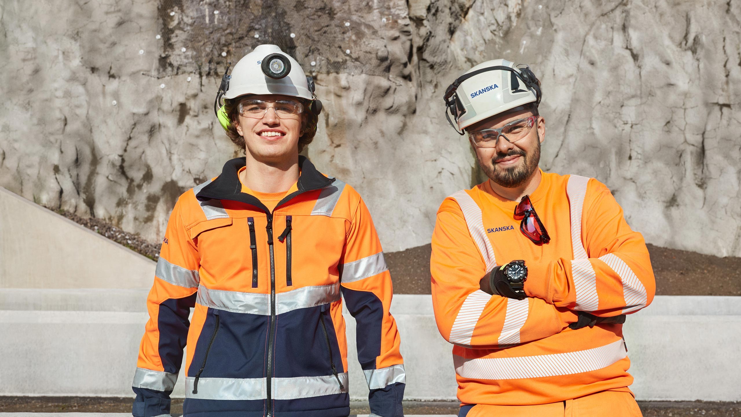 Construction workers in safety gear at rocky site outdoors Two construction workers wearing safety gear and helmets standing in front of a rock wall.