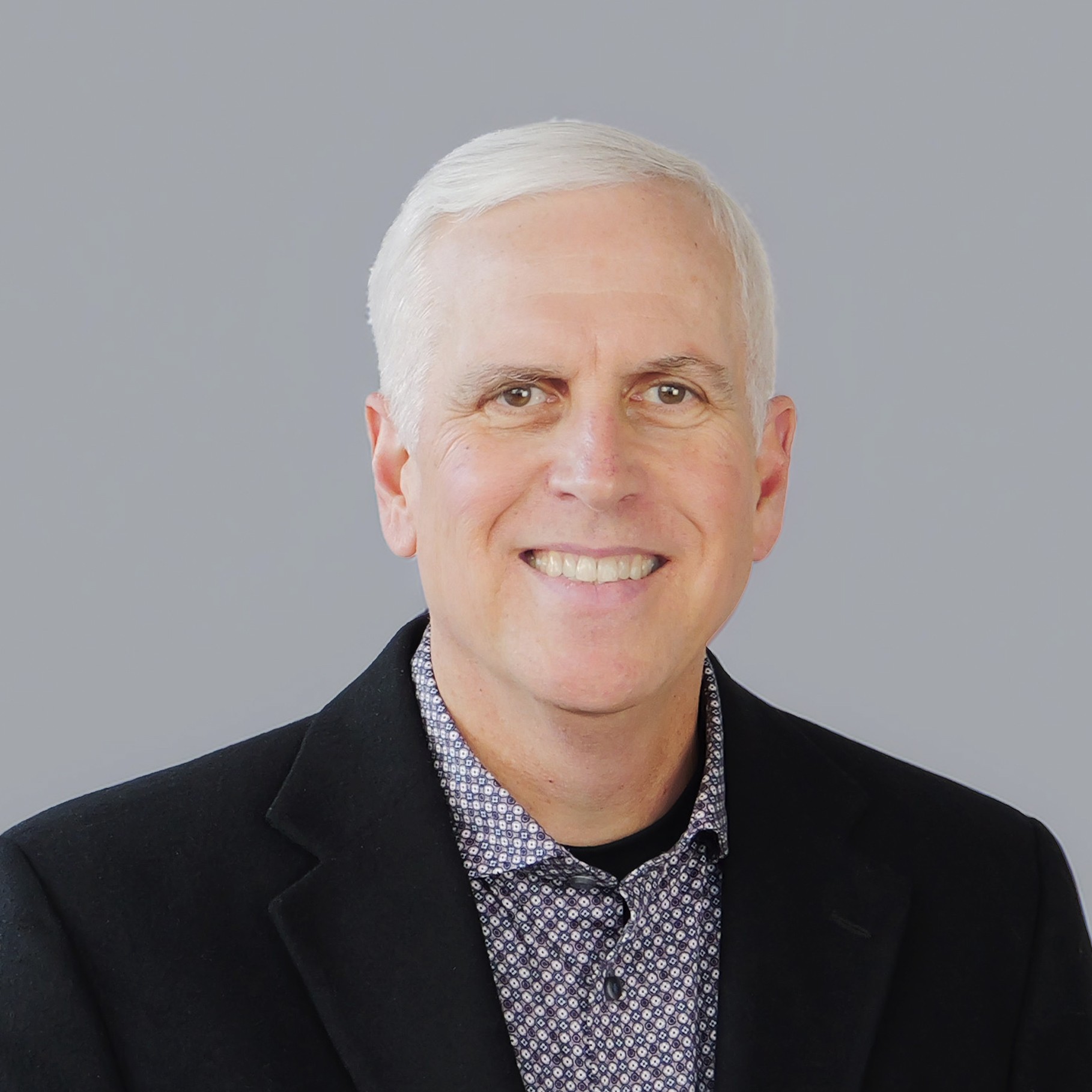 A man with white hair wears a patterned shirt and black blazer against a gray background.