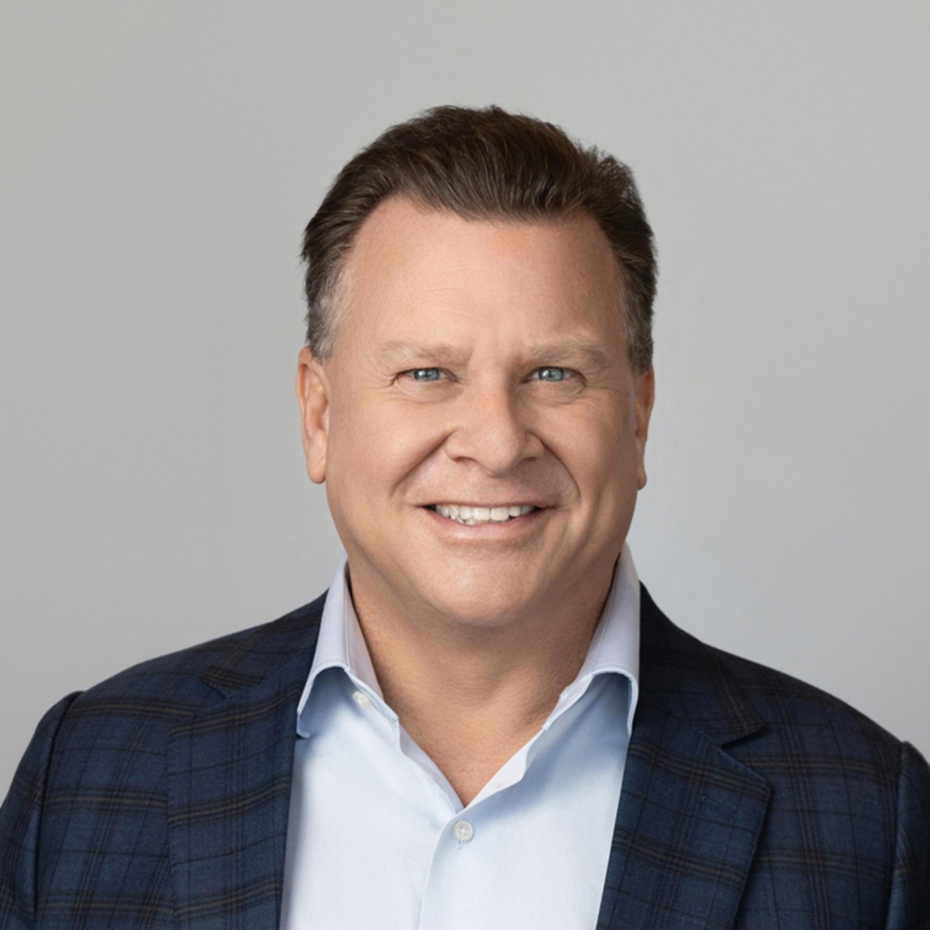 Man in a plaid suit jacket and light blue shirt against a neutral background.