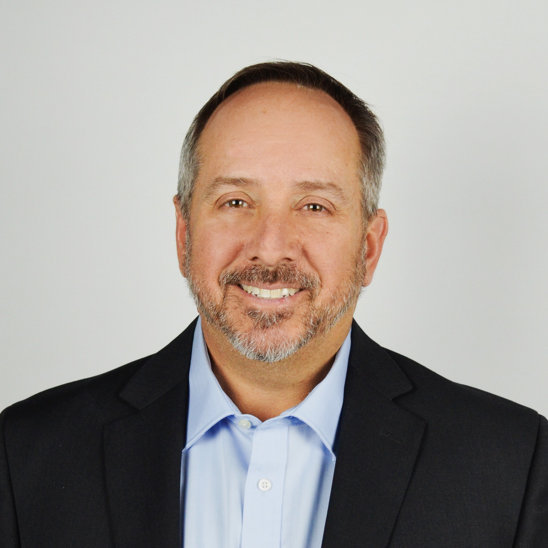 Professional Man in Suit and Tie Against Plain Background A man in a black suit and light blue shirt stands against a neutral background.