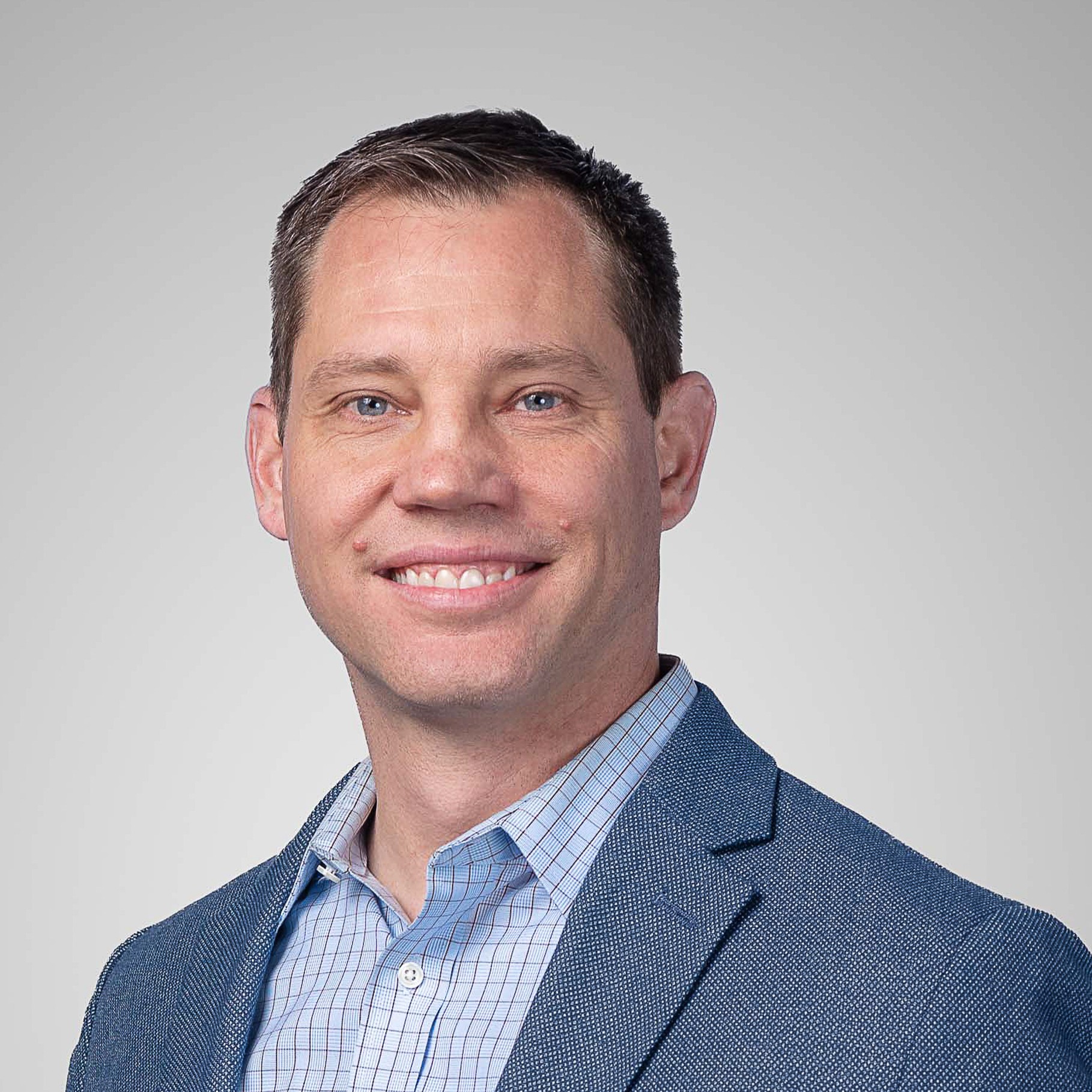 A man in a blue blazer and checkered shirt against a gray background.