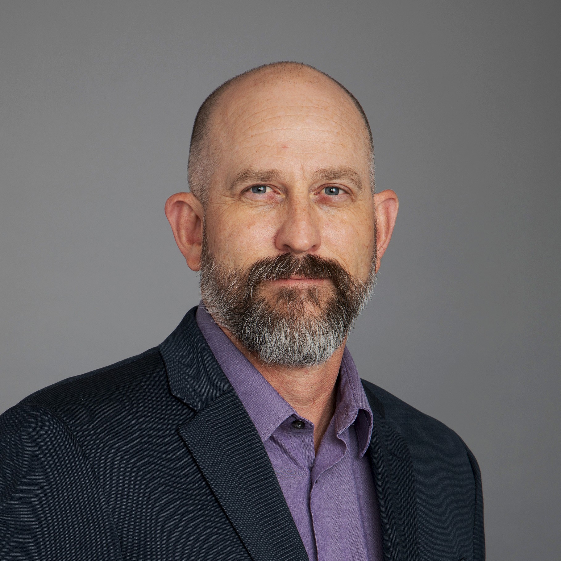 Clark Peterson in Suit and Purple Shirt on Grey Background Clark in a suit jacket and purple shirt, standing against a neutral gray background.