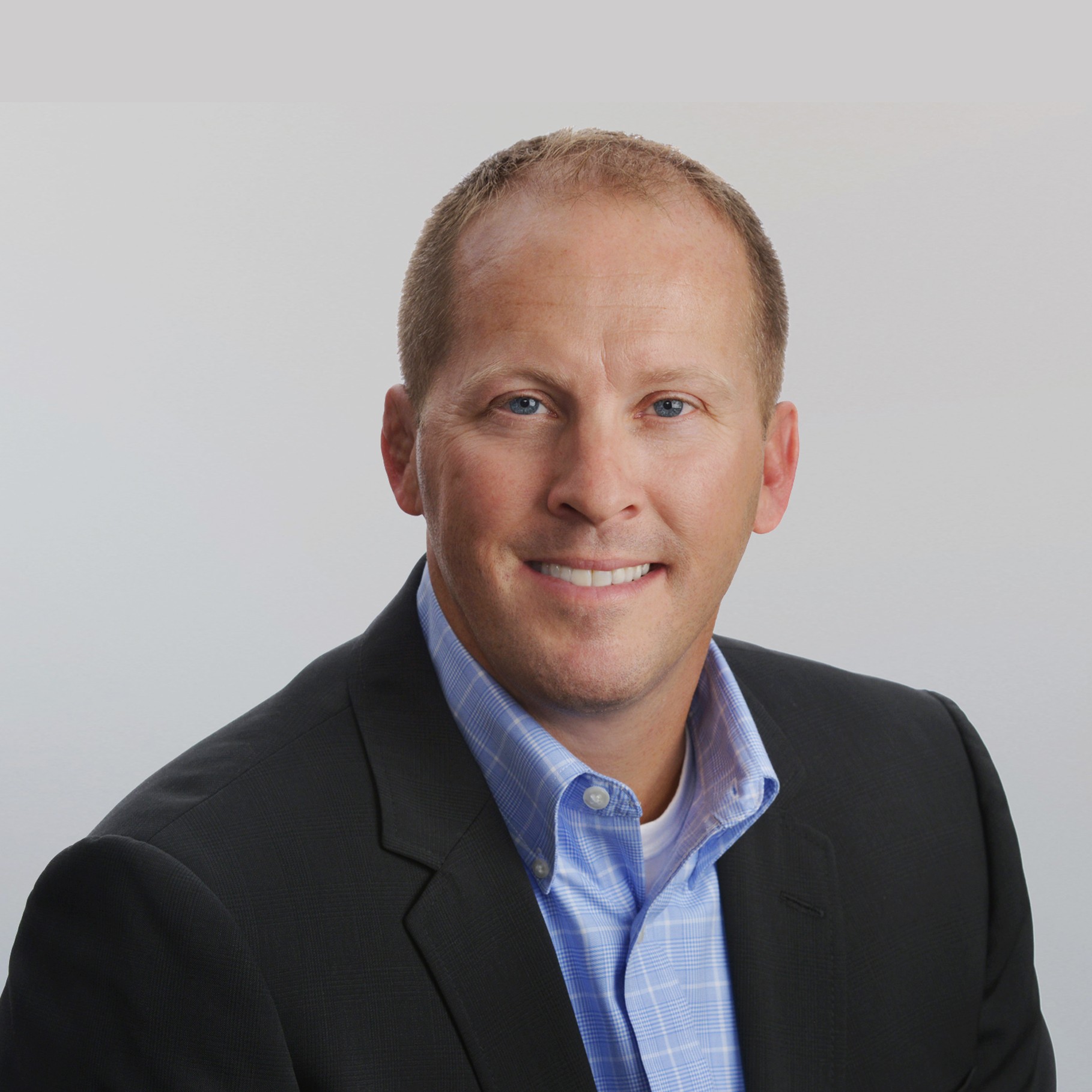 A man in a black suit and blue checkered shirt sits against a light gray background.