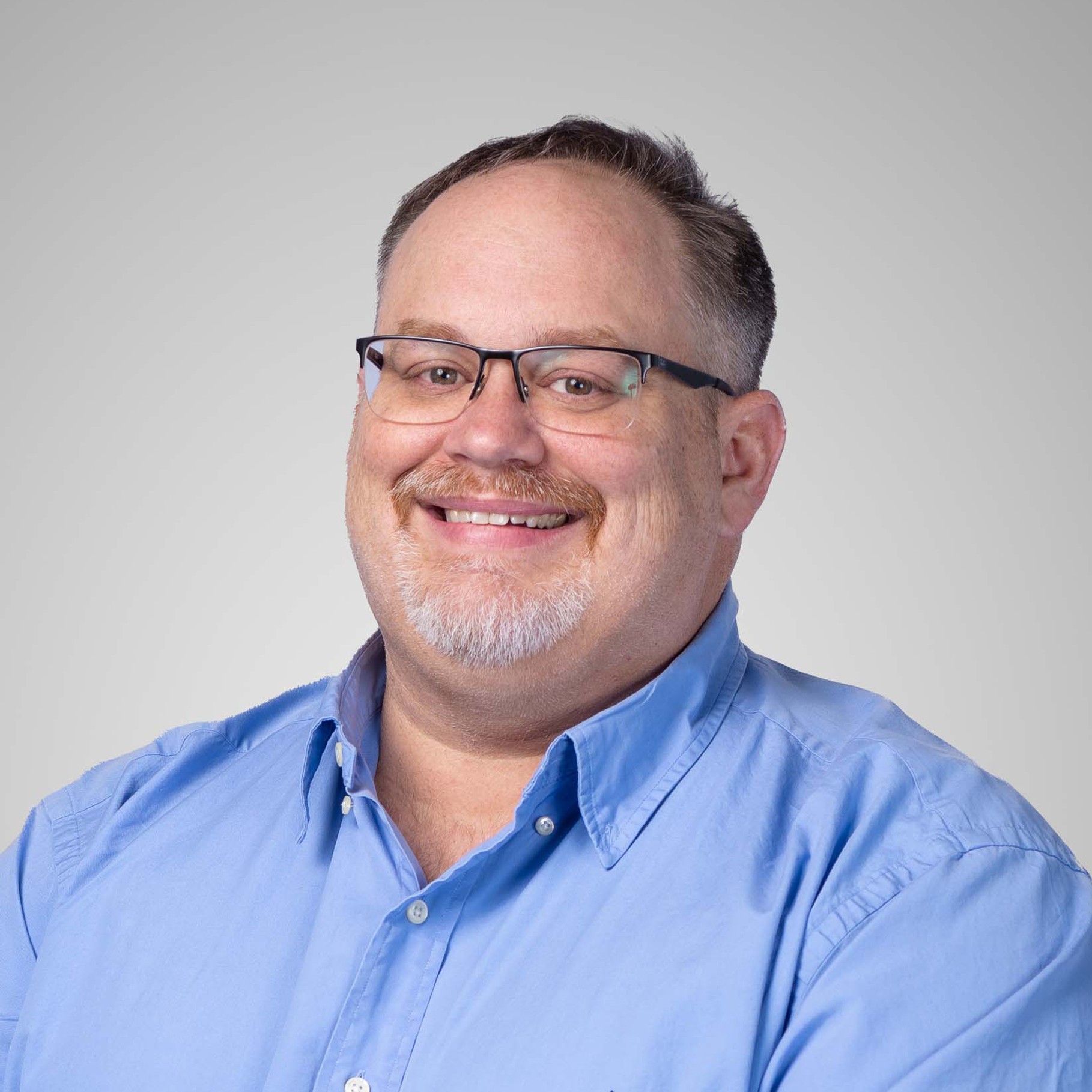 Man in Blue Shirt Against Gray Background A man wearing a blue button-up shirt with crossed arms, set against a neutral gray background.