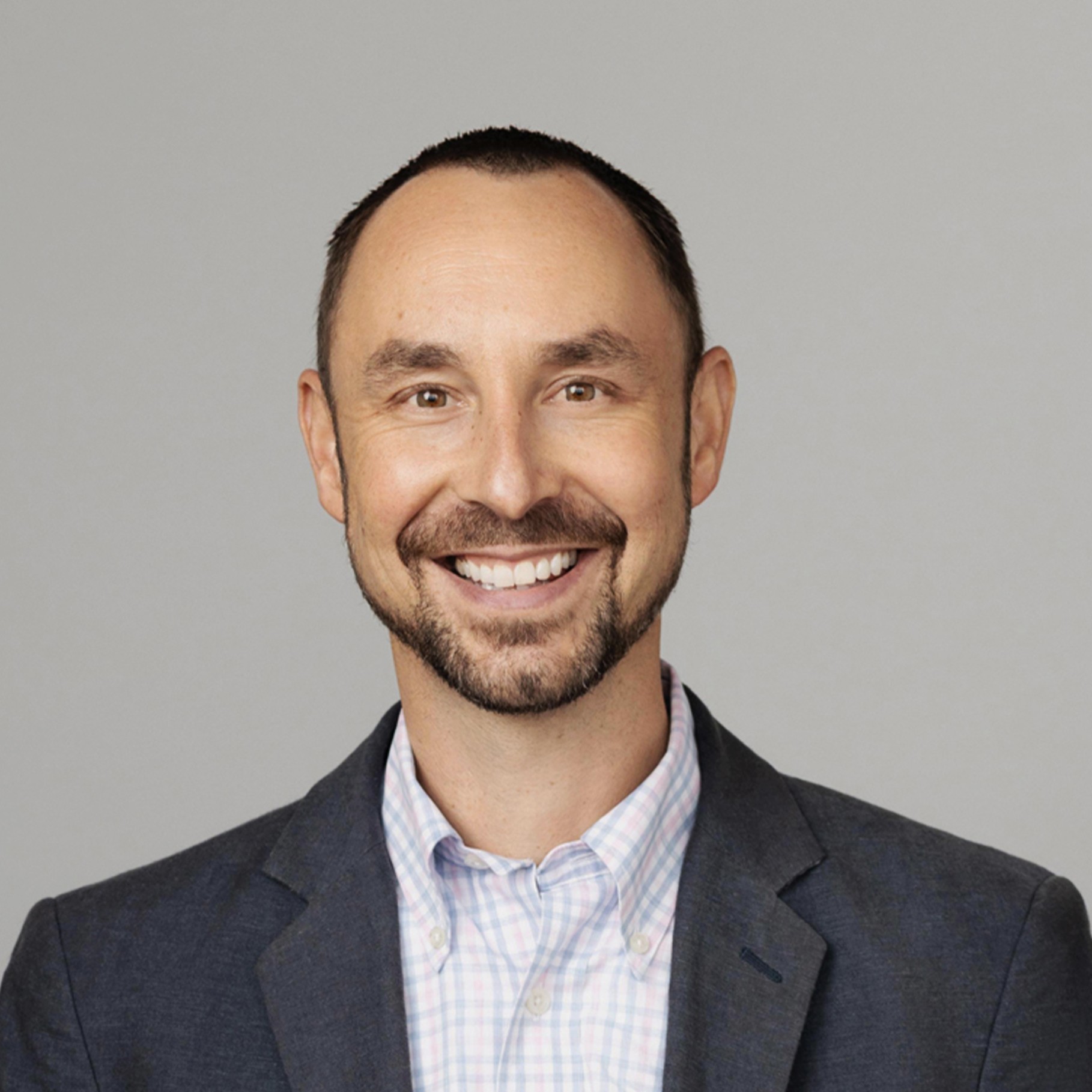 Man in a dark blazer and checkered shirt against a plain gray background.