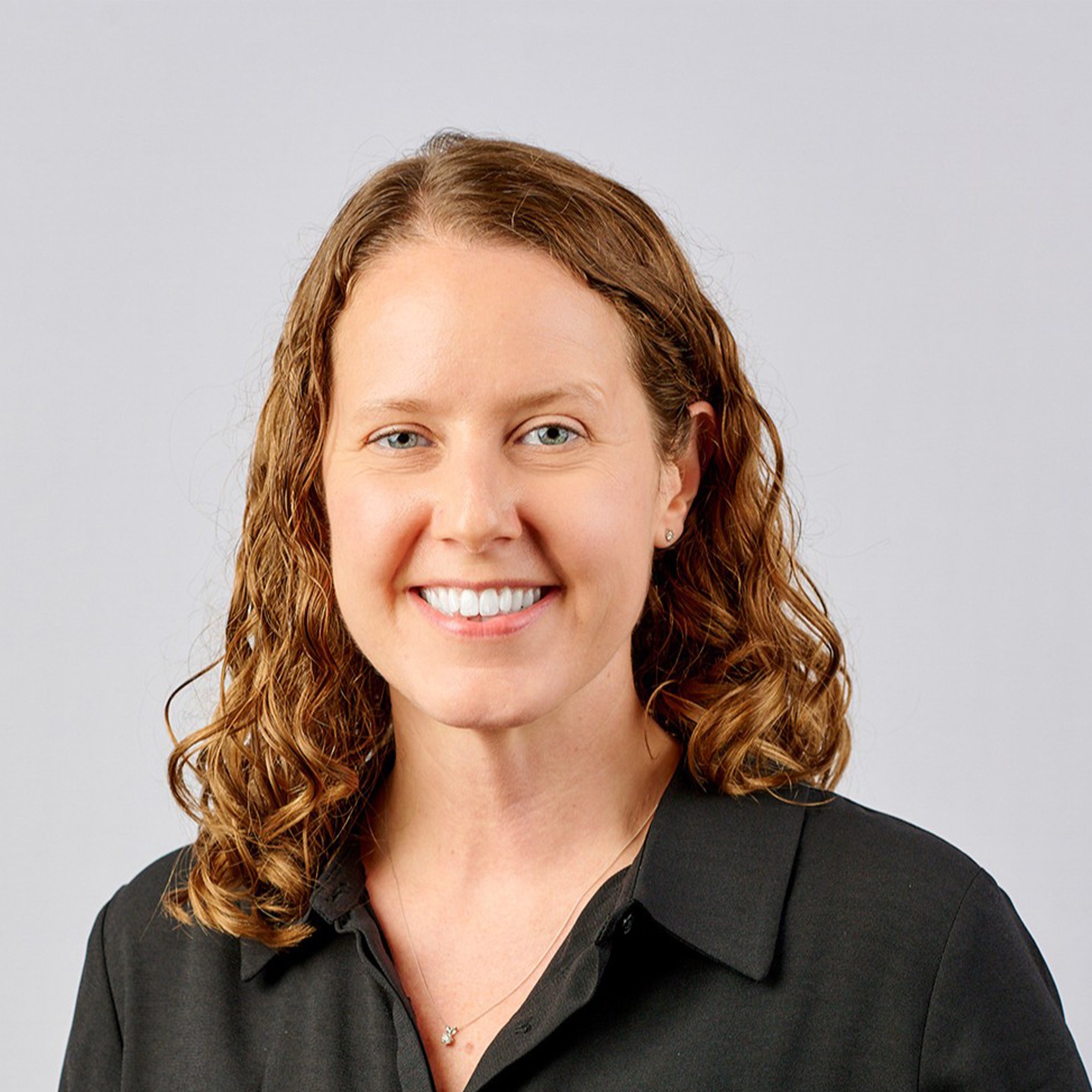 A person with curly, brown hair wearing a dark collared shirt against a plain background.