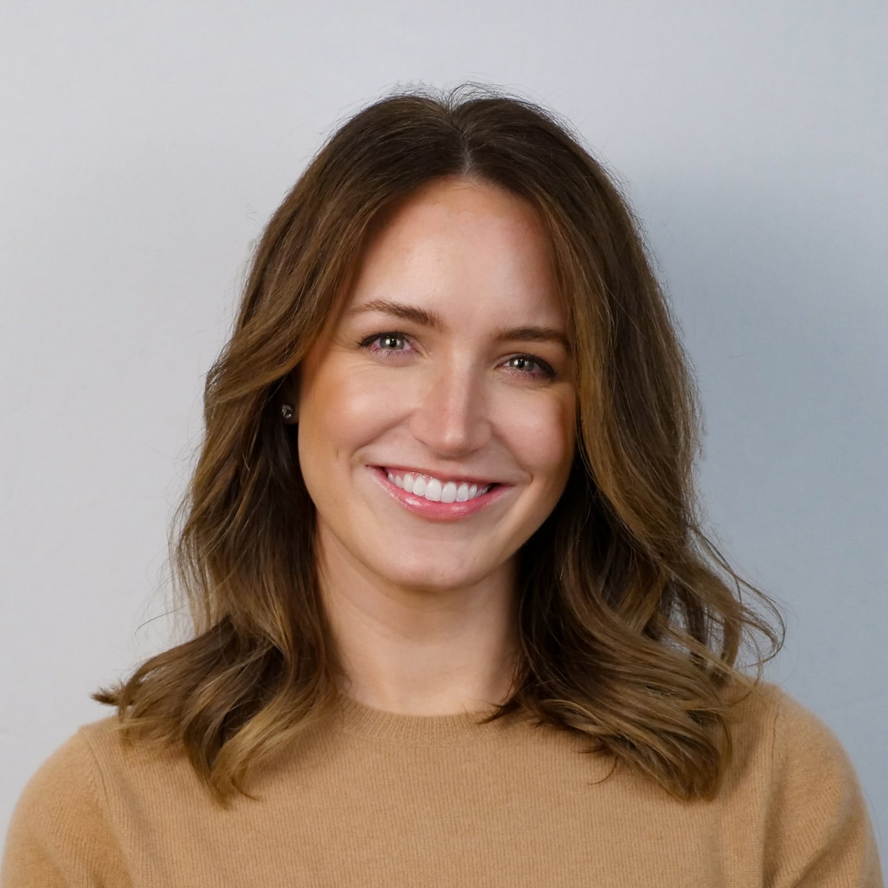 Woman with wavy brown hair wearing a beige sweater against a light gray background.