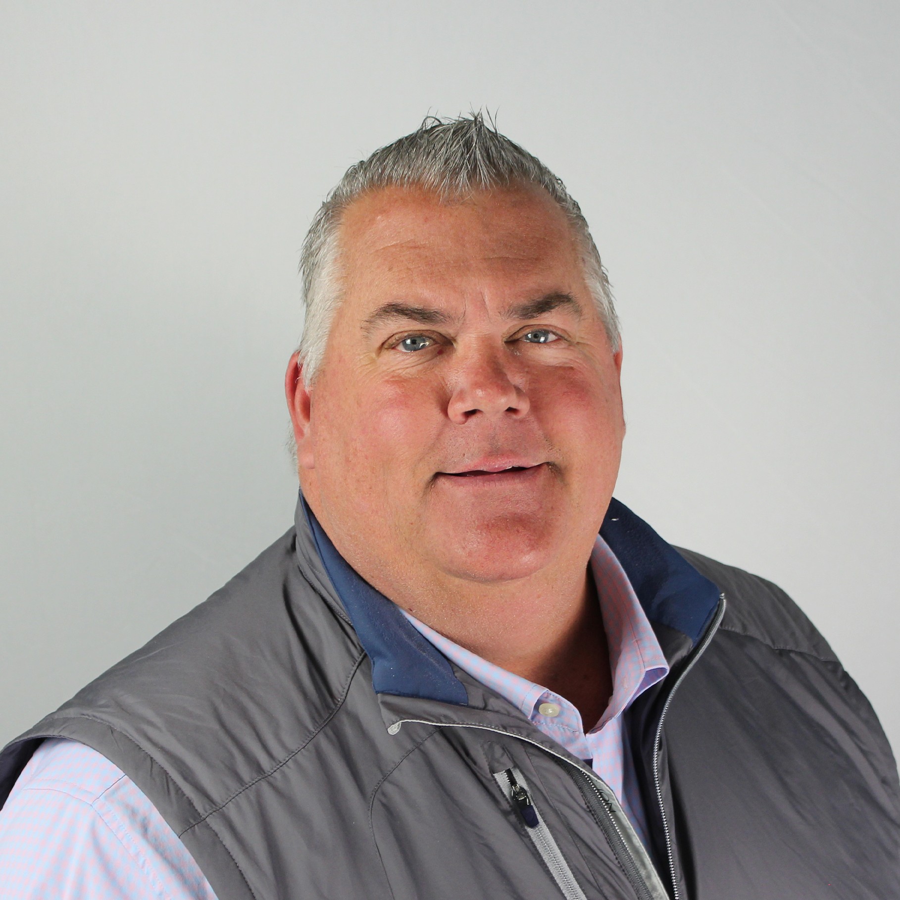A man wearing a gray vest over a light-colored shirt against a plain background.