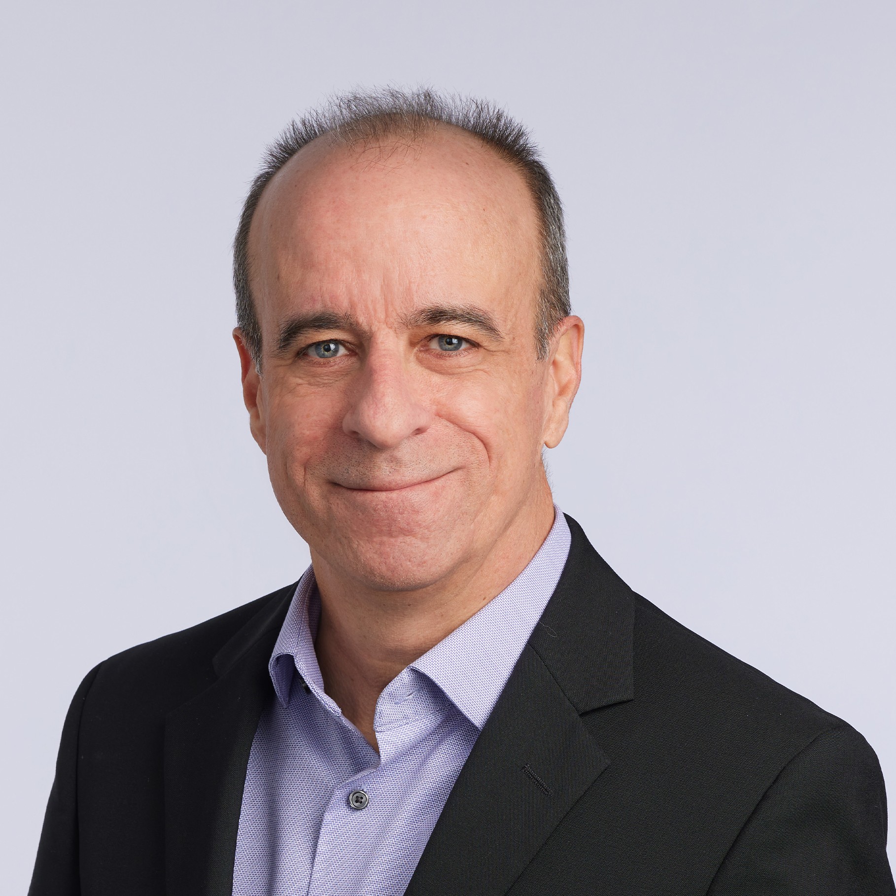 Man in a black blazer and light purple shirt against a plain background.