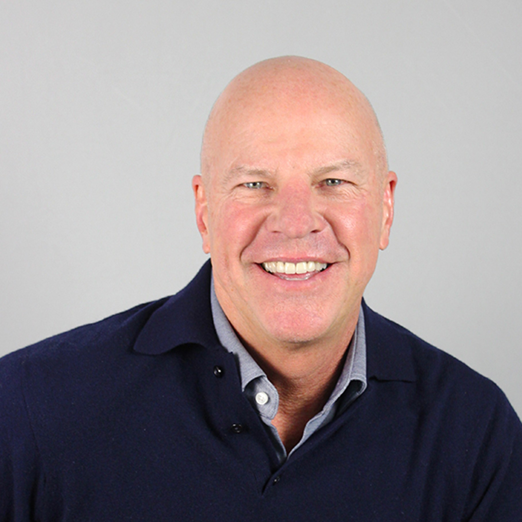 Man in Blue Jacket Posing Against a Neutral Background A person in a dark sweater over a collared shirt against a plain gray background.