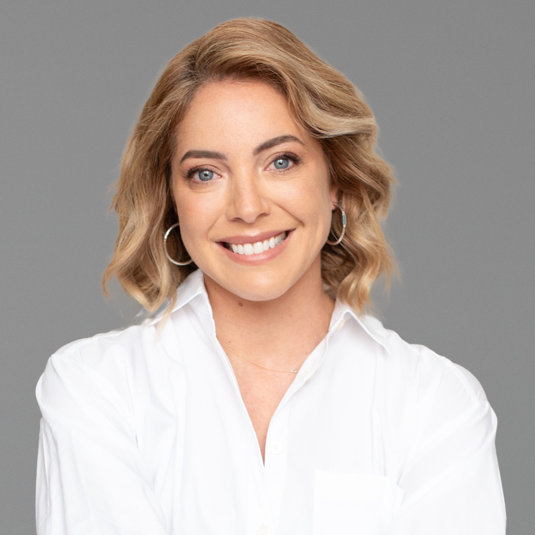 Stylish Woman in White Shirt with Wavy Blonde Hair A person with wavy blonde hair wearing hoop earrings and a white shirt against a beige background.