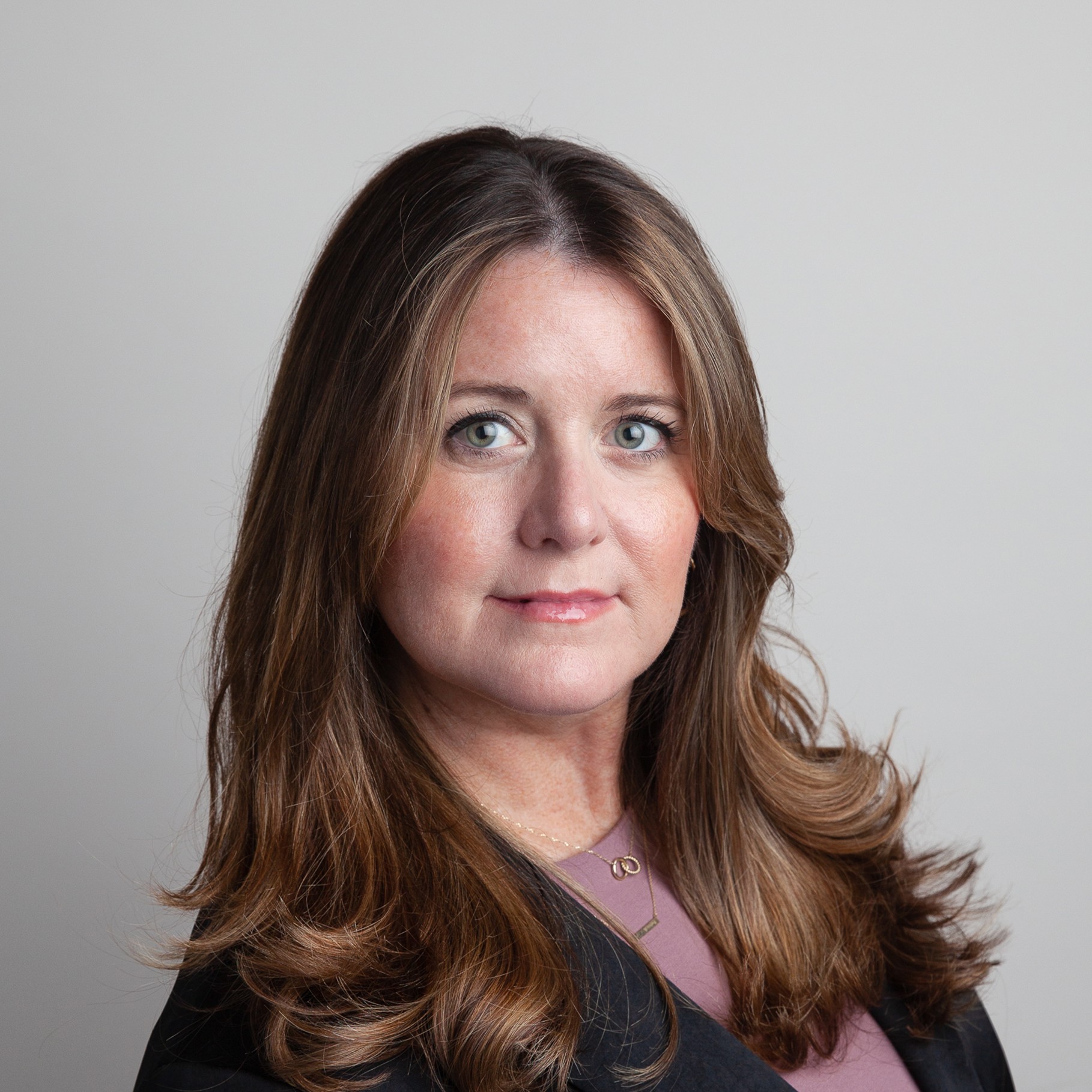 A woman with long, wavy brown hair wearing a black blazer and mauve top, posing confidently.