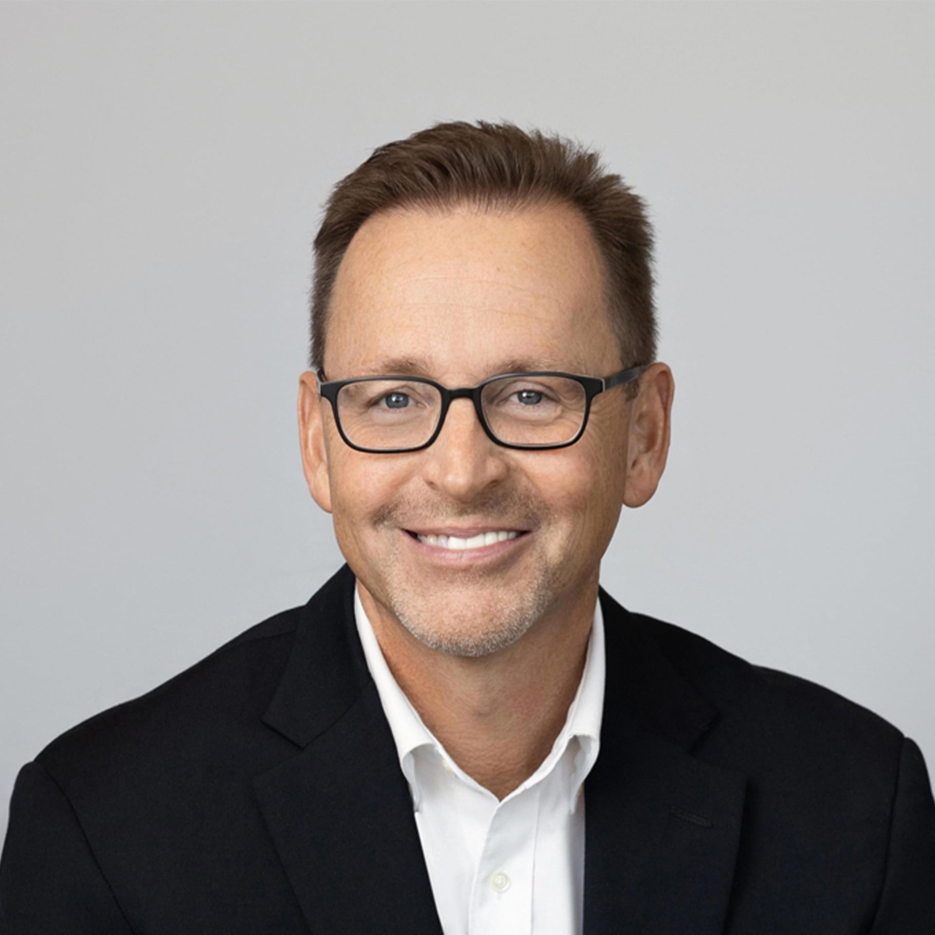A man in a black suit jacket and white shirt sits against a plain gray background.