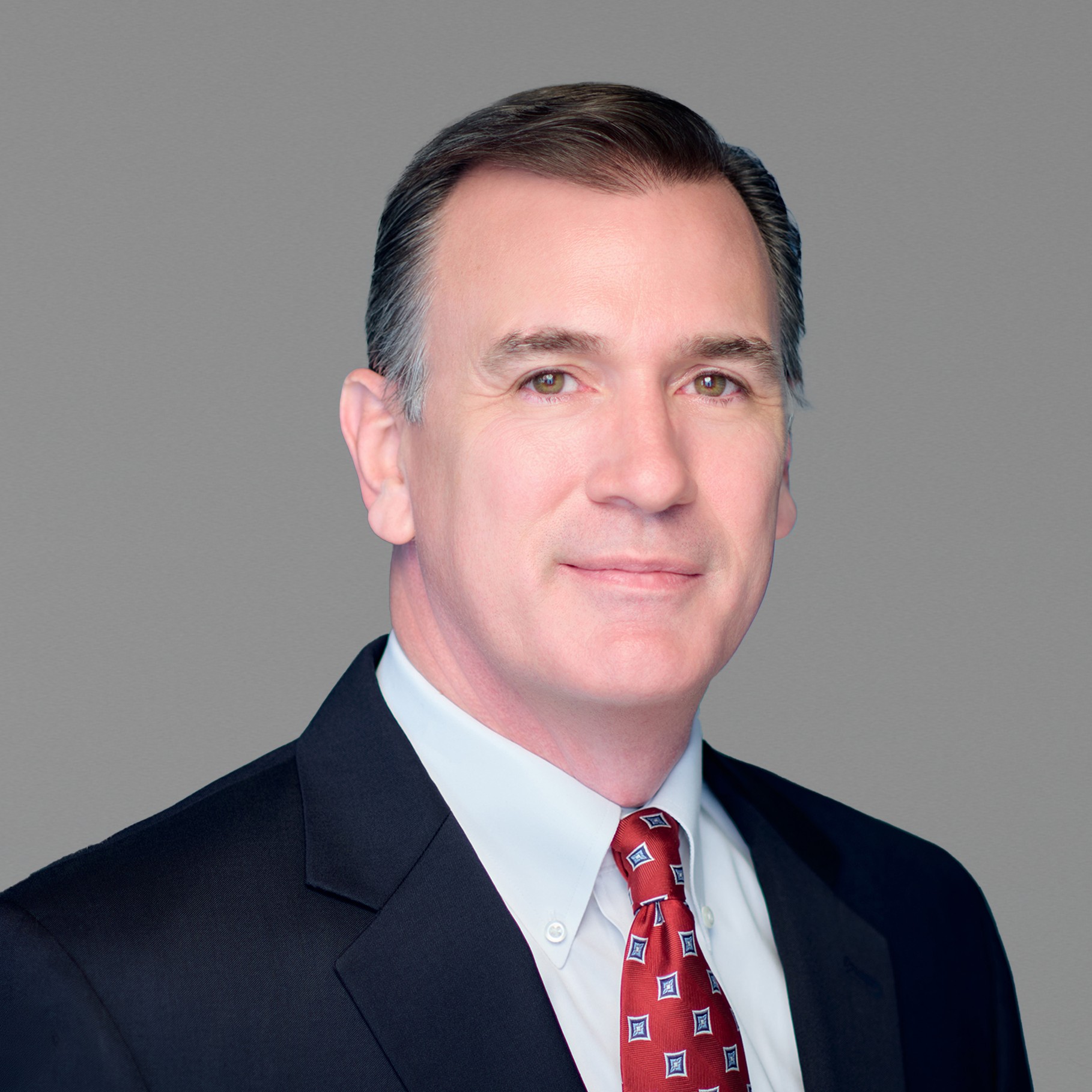 Professional businessman in suit with red patterned tie, gray background Mark wearing a dark suit and red patterned tie against a gray background.
