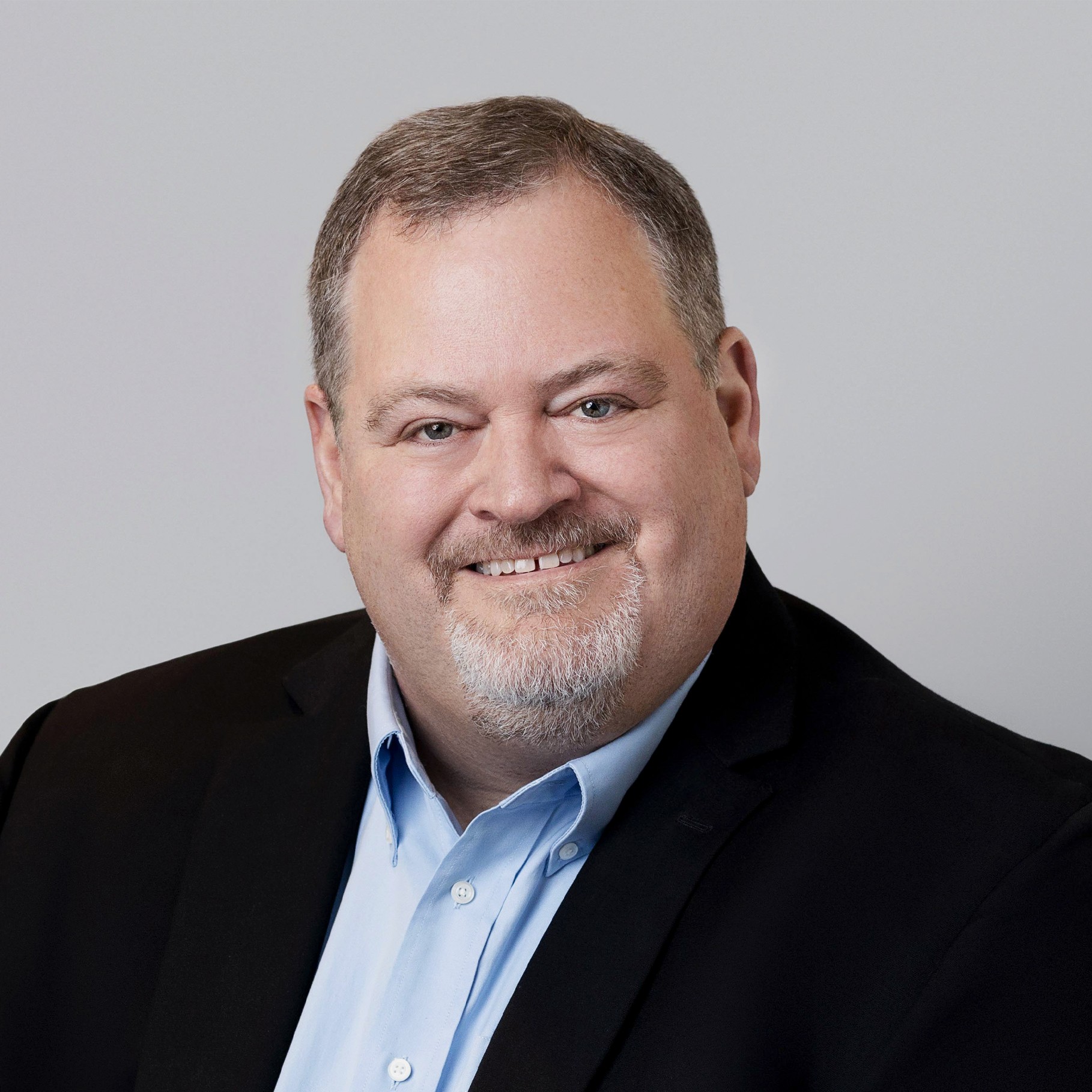 A person in a black blazer and blue shirt against a neutral gray background.