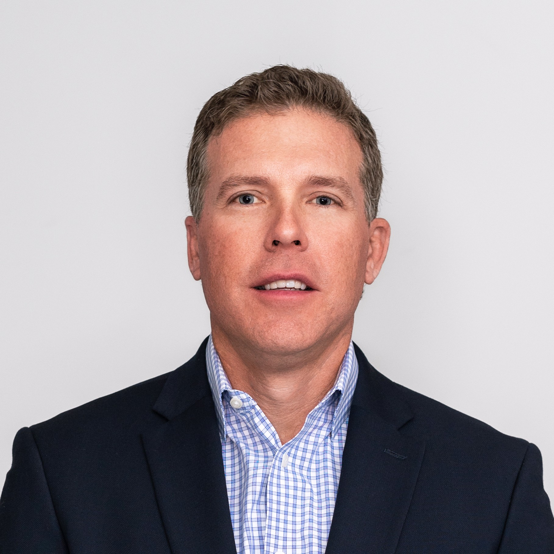 A man in a navy suit and checked shirt stands against a plain white background.