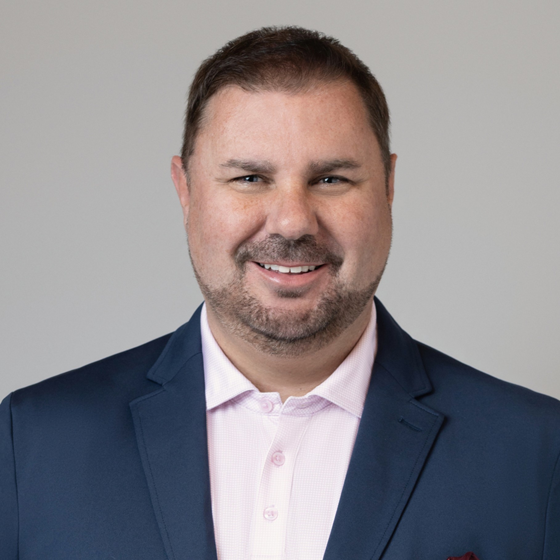 Professional businessman in suit with gray background portrait Stephen wearing a navy blazer and light pink shirt against a plain gray background.