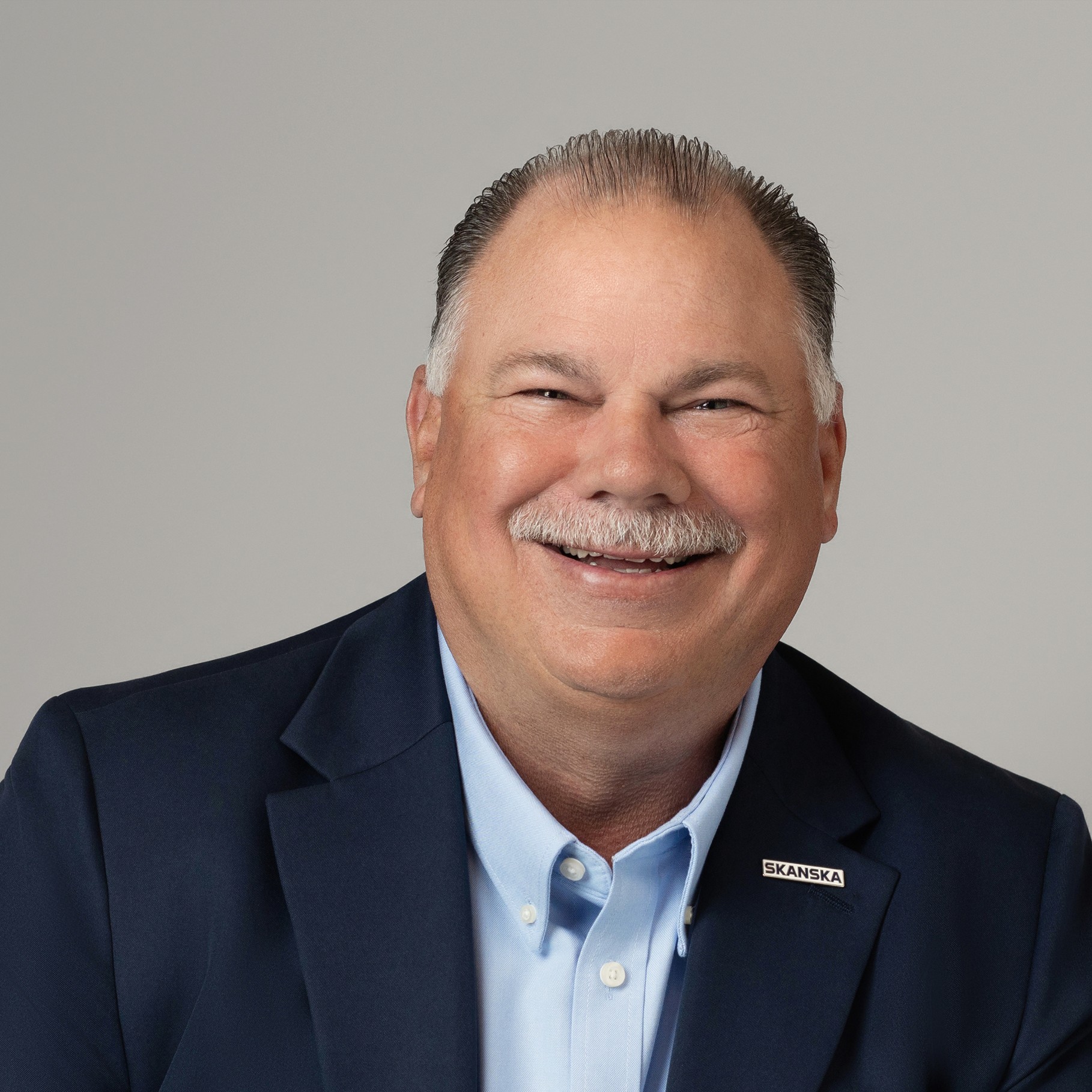 Middle-aged man in checkered shirt with neutral gray background Steve wearing a navy suit and light blue shirt with a Skanska lapel pin on a gray background.